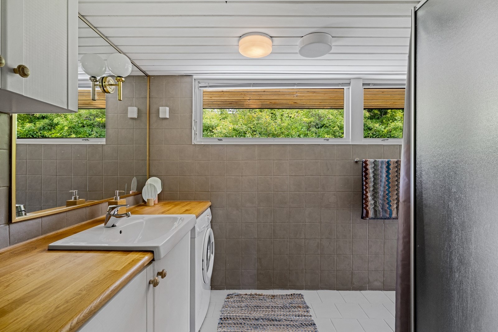 Badet har grå fliser på veggene, hvitmalt panel i himlingen og lyse fliser på gulv med gulvvarme. Galleribilde