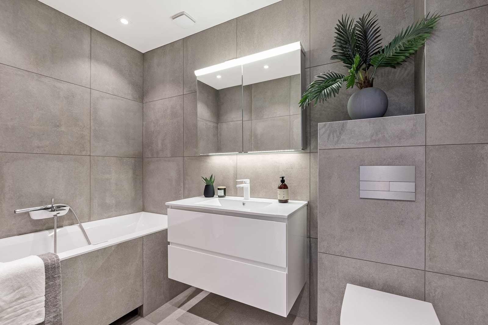 Innbydende og lekkert bad med moderne flisvalg og varme i gulv. Badet er utstyrt med plassbygd badekar, hvit skuffeinnredning, speilskap på vegg og vegghengt toalett. Galleribilde