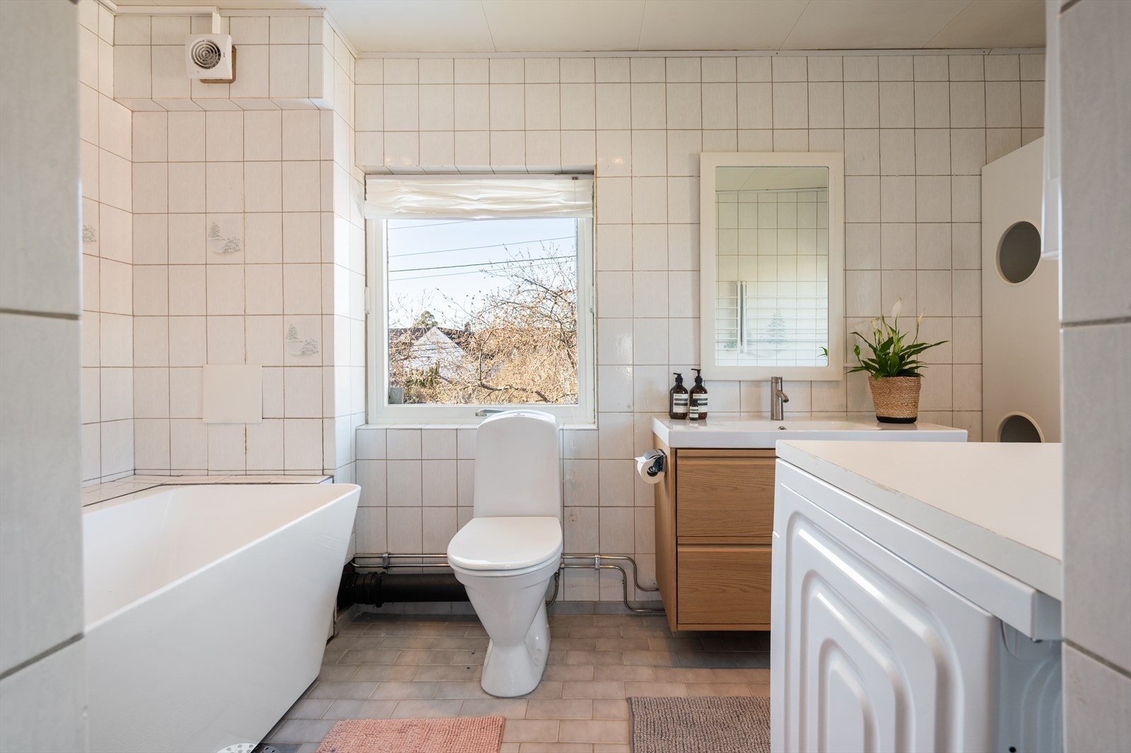 Romslig, eldre baderom med stort vindu som gir gode, naturlige lysforhold og gode luftemuligheter. Badet er innredet med gulvstående toalett, servant nedfelt i baderomsinnredning, opplegg for vaskemaskin og tørketrommel samt badekar. Galleribilde
