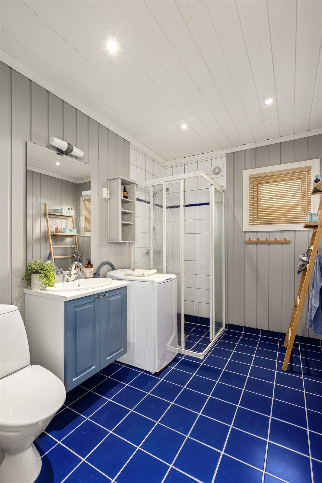 Badet har flislagt gulv med varmekabler og malt panel på vegger (flis i dusjsone). Rommet inneholder servant med skap, dusjhjørne, wc og opplegg for vaskemaskin. Galleribilde