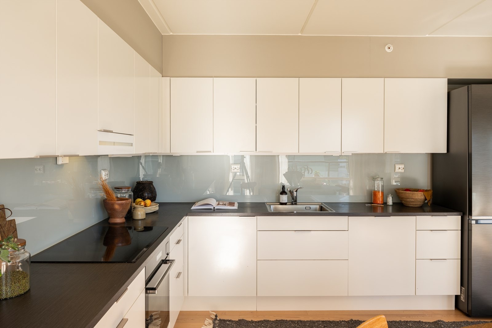 Kjøkkenet er utformet i L-form med hvit, glatt innredning, mørk laminatbenkeplate med stålvask og elegante, spesialtilpassede glassfelt over hele benken. Galleribilde