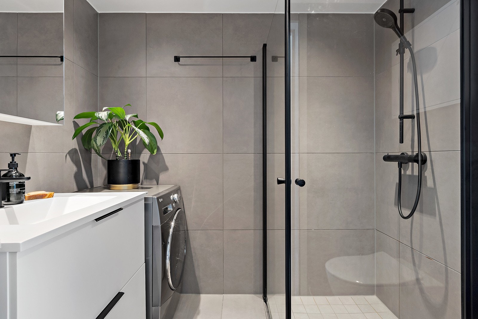 Badet har flislagt gulv med gulvvarme, fliser på vegger og malt innvendig tak. Baderomsinnredning med heldekkende servant og underskap med skuffer, speilskap med overlys, veggmontert toalett og dusjhjørne m/ svingbare glassdører og opplegg for vaskemaskin Galleribilde