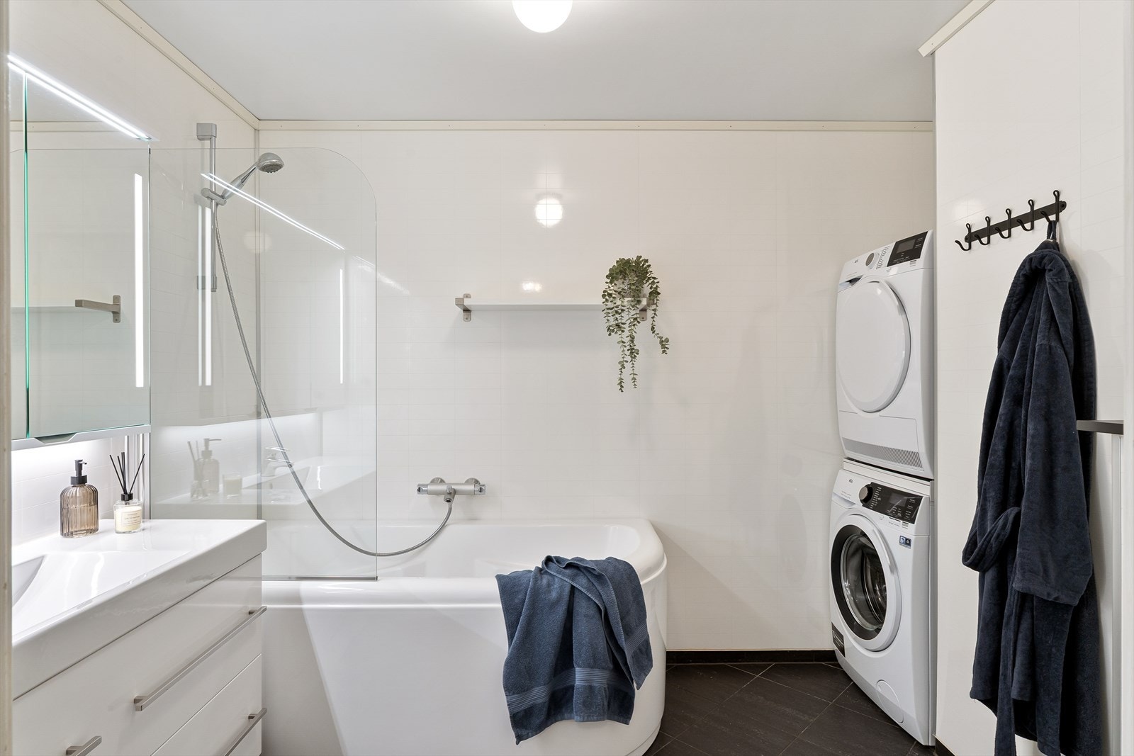 Romslig bad som er utstyrt med baderomsinnredning med nefelt vask, speilskap, badkar med dusjvegg, vegghengt toalett og opplegg/plass for vaskemaskin og kondenstørketrommel. Galleribilde