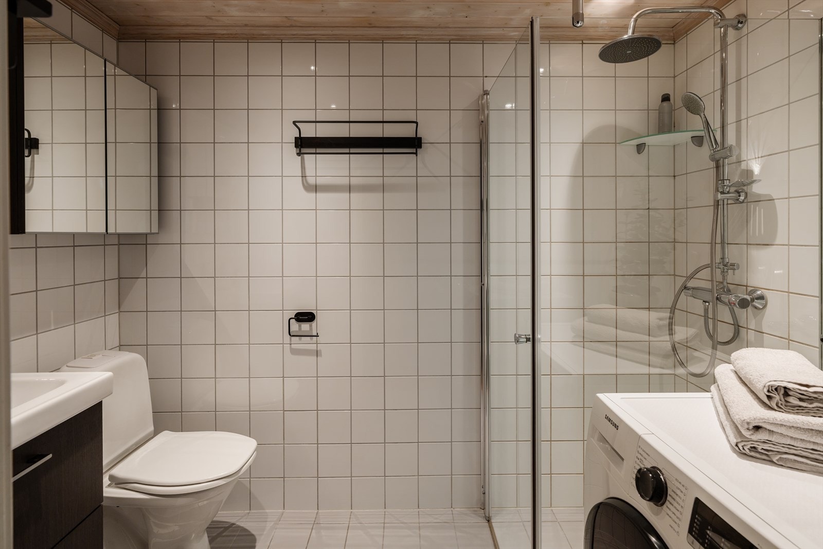 Badet er innredet med klosett, dusjhjørne, servant med speil og underskuffer, samt håndkleoppheng på vegg. Galleribilde