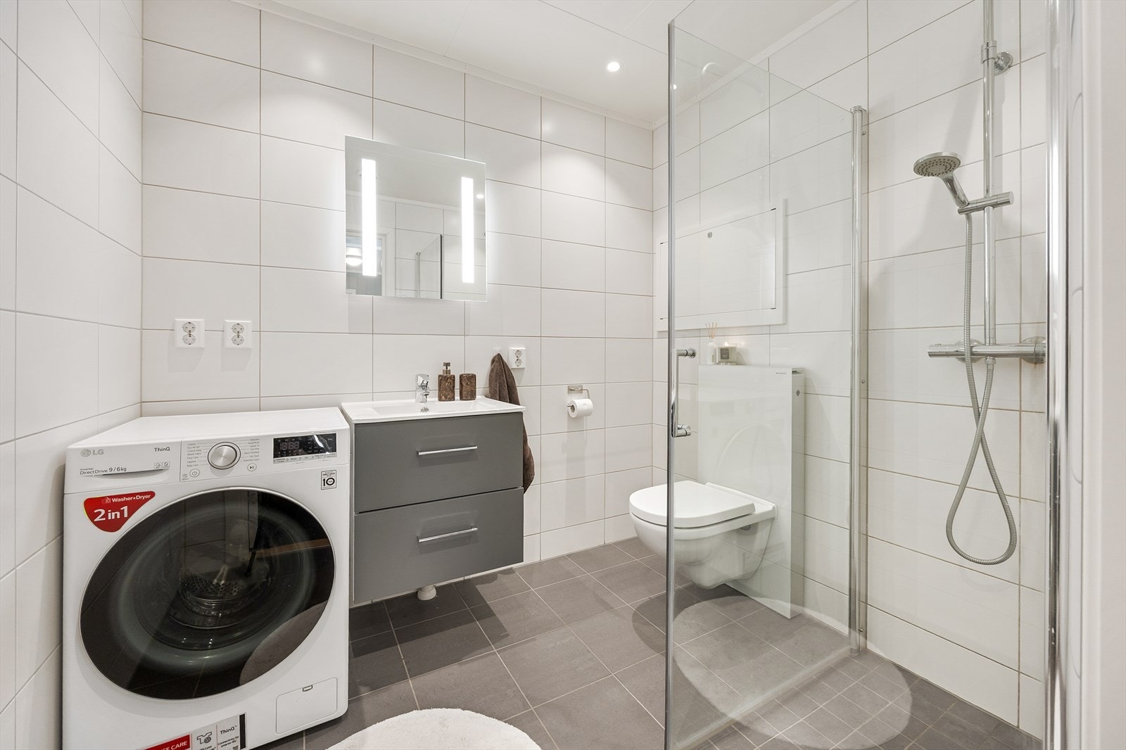 Badet er innredet med vegghengt toalett, servant i baderomsinnredning, speil med integrert belysning og dusjhjørne med innfellbare glassvegger. Galleribilde