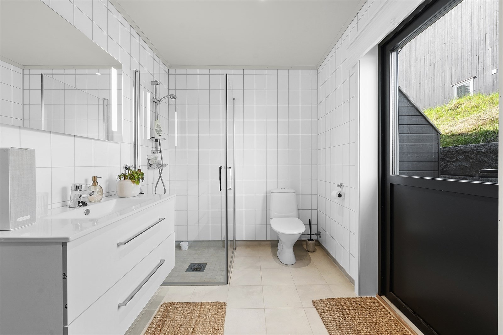 Badet er utstyrt med heldekkende servant over skuffeinnredning, speil med belysning, dusjhjørne med innfellbare glassvegger og toalett. Flislagt gulv med varmekabler og baderomsplater på vegger. Dør ut Galleribilde