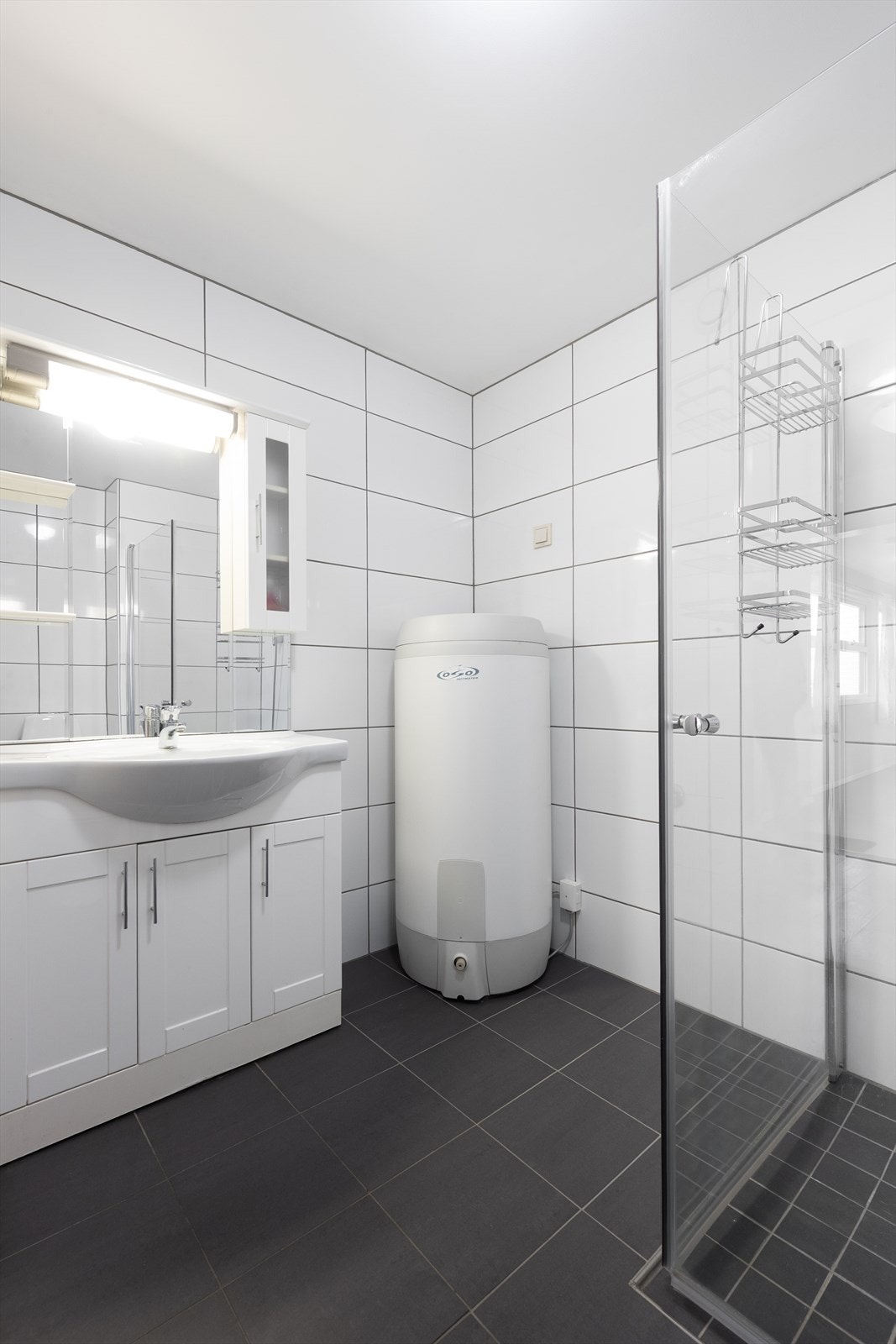 Badet inneholder heldekkende servant med underskap, dusjhjørne med innfellbare glassdører, gulvmontert toalett, varmtvannstank og opplegg til vaskemaskin. Galleribilde