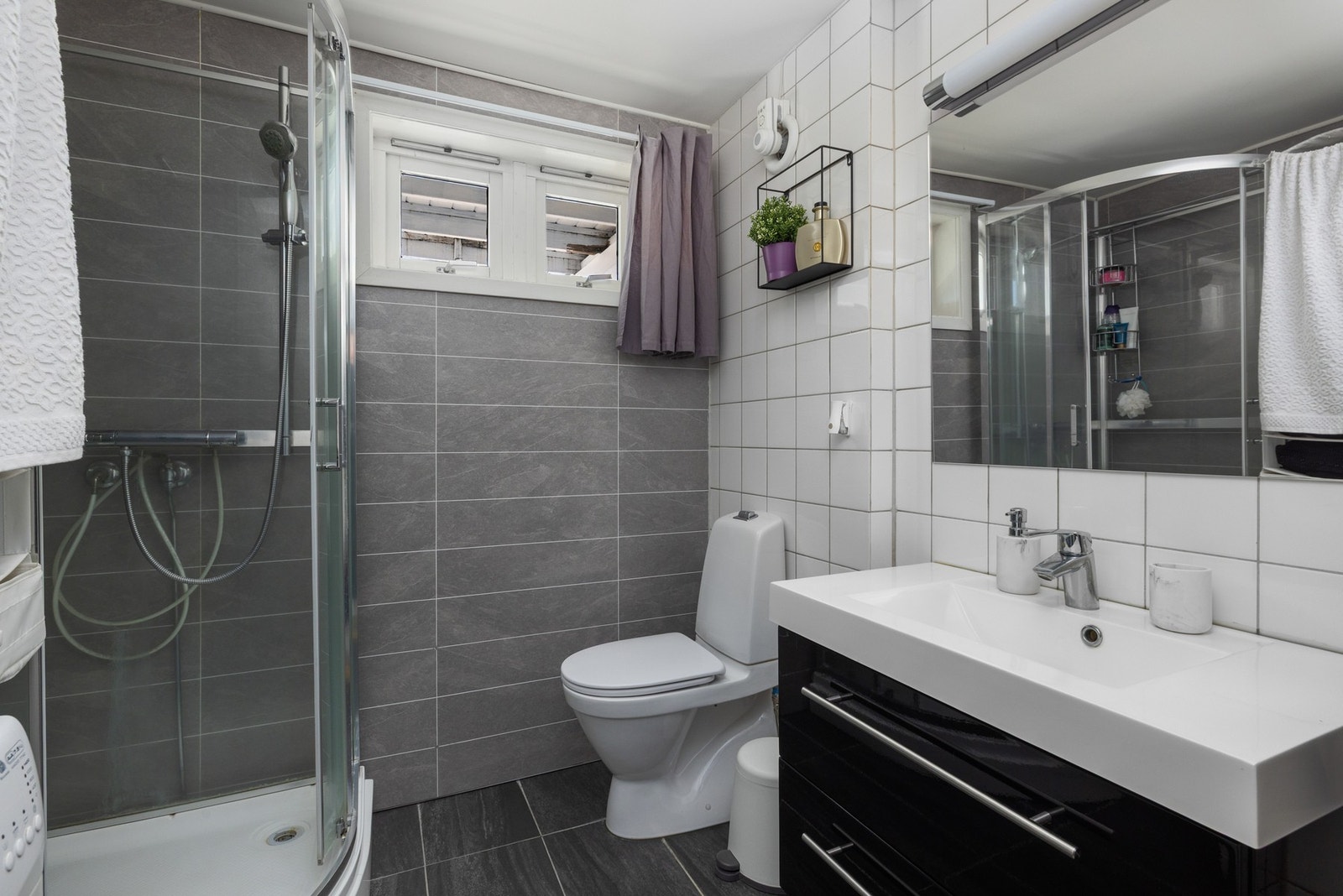 Badet har dusjkabinett, toalett, og servant med underskap. Lyse fliser på vegger og mørke fliser på gulv. (utleie) Galleribilde