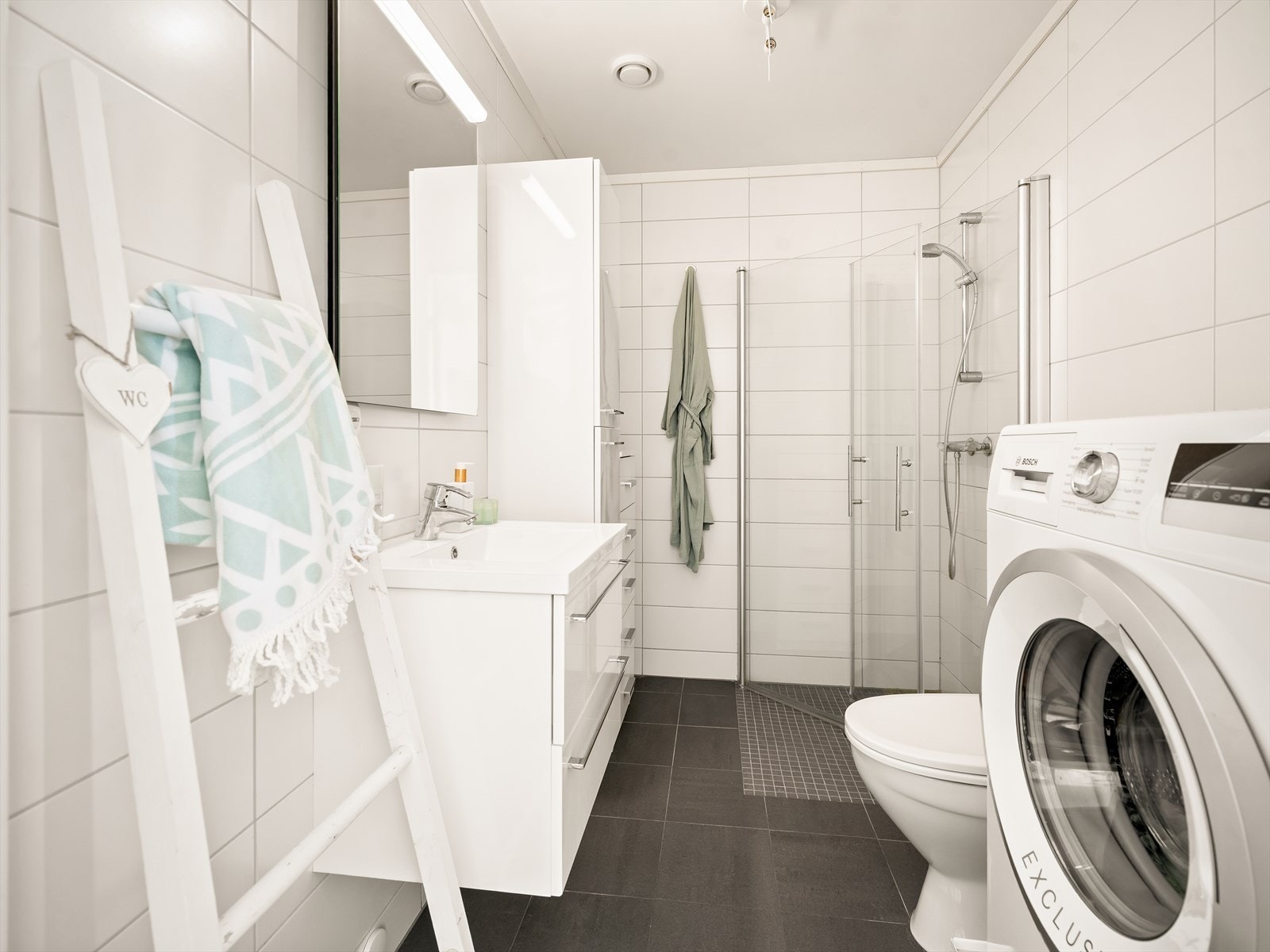 Badet er innredet med servantskap med nedfelt servant, høyskap, gulvmontert toalett, dusjhjørne med dusjvegger og opplegg for vaskemaskin. Galleribilde