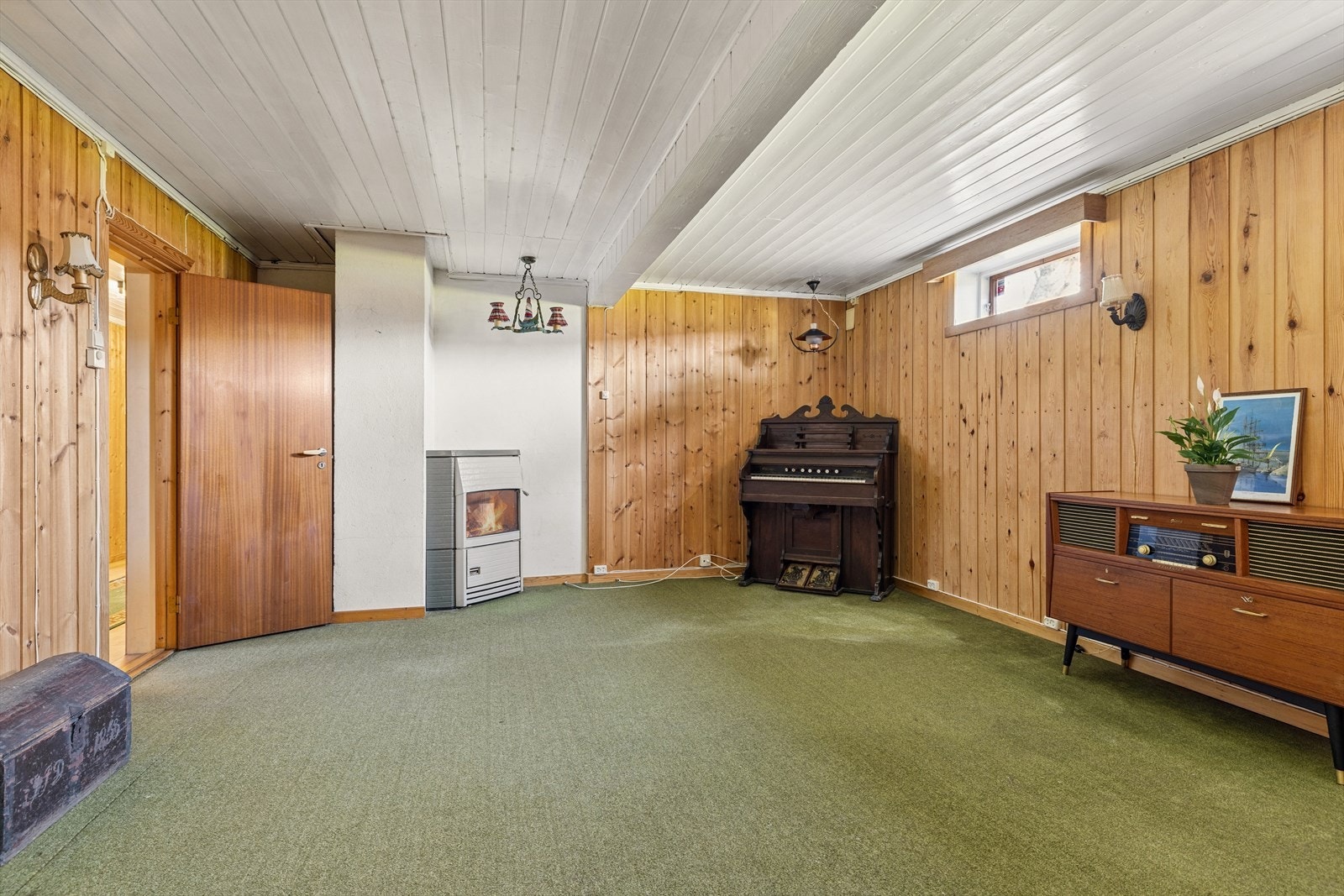 Kjellerstue med teppe på gulvet og trepanel på vegger og i tak. Rommet er ikke godkjent for varig opphold. Galleribilde