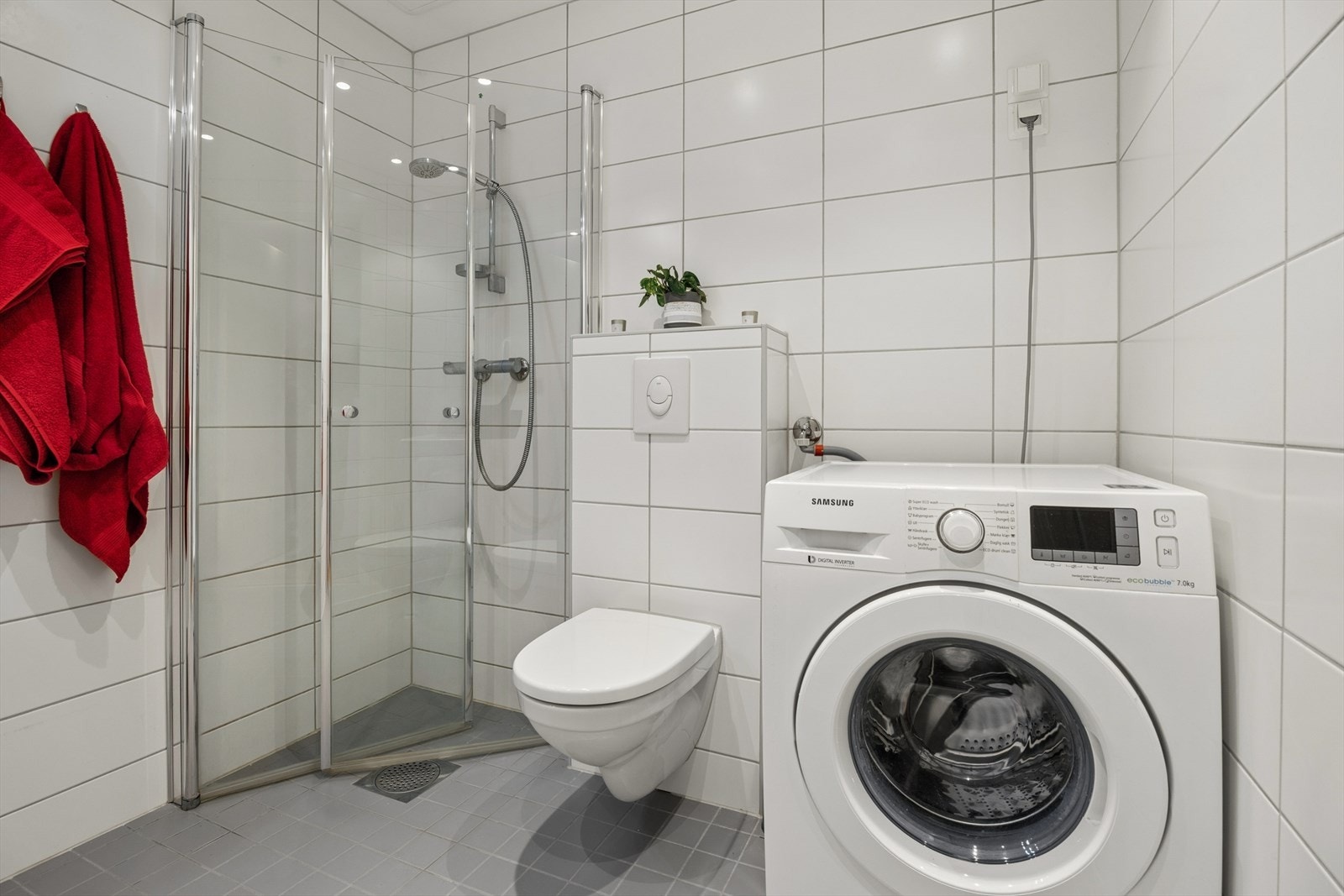 Badet er utstyrt med vegghengt toalett, dusjhjørne med innsvingbare glassdører, innredning med servant, speilskap og opplegg for vaskemaskin. Galleribilde