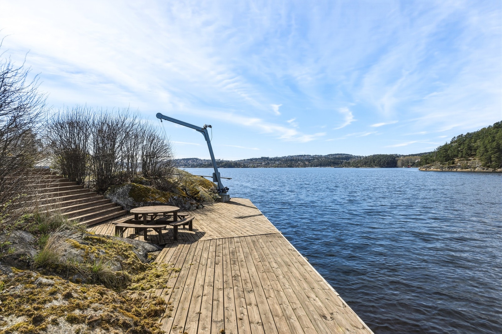 Brygge med trapp, kran og sittegruppe ved vannet, omgitt av naturlig vegetasjon og utsikt over fjorden. Galleribilde