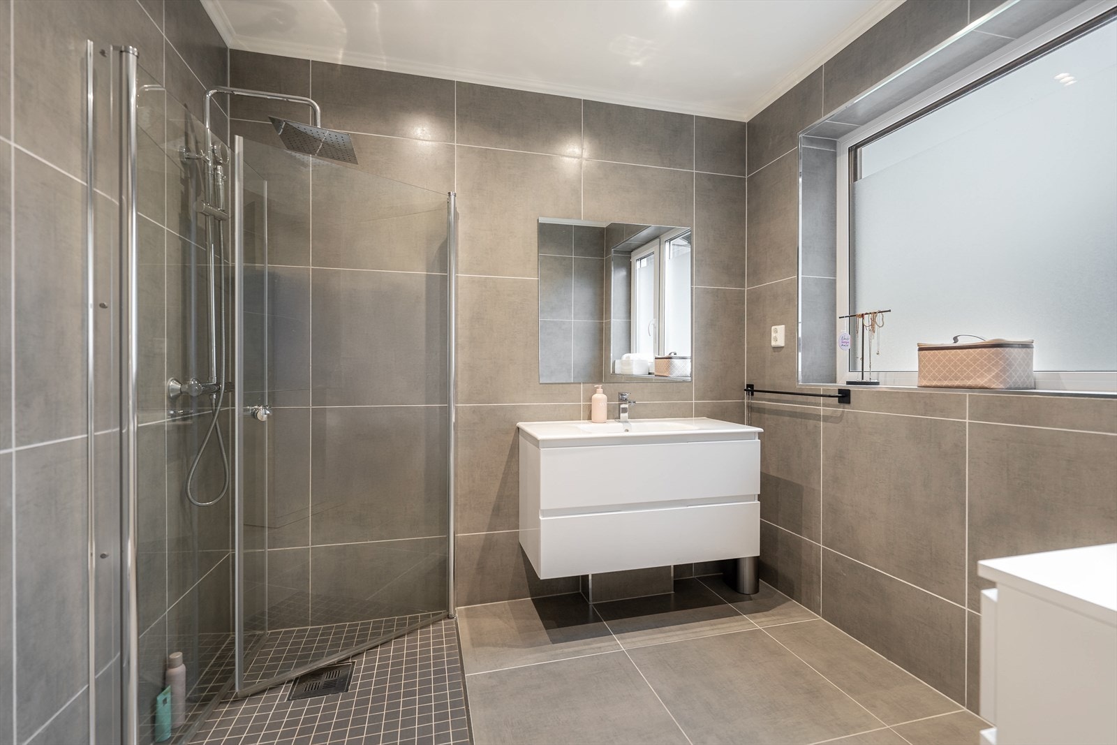 Fra entreen er det adkomst til ett av boligens bad. Badet i 1.etasje er flislagt med varmekabler i gulvet og malte slette flater i taket. Badet er innredet med nedfelt servant, veggmontert toalett og dusjhjørne med praktiske dusjdører. Galleribilde