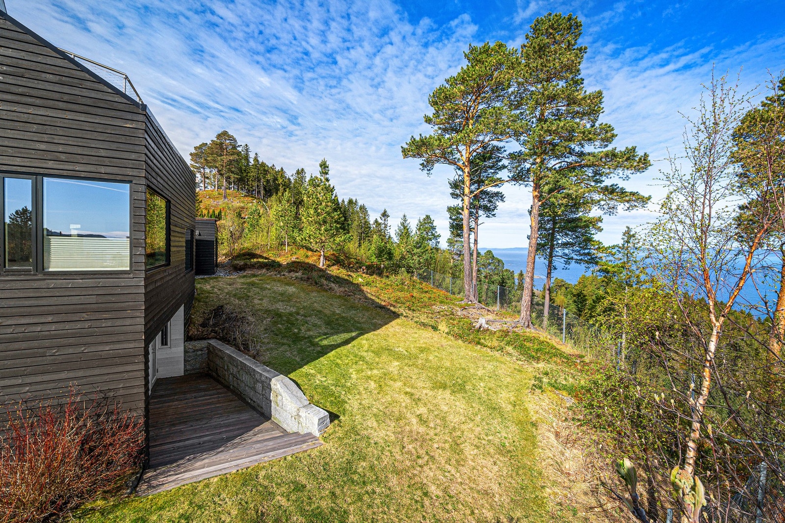 Hagen fortsetter rundt huset, ut mot utsiktssiden. Perfekt for barna å kunne løpe rundt huset. Tomta grenser ut mot blåbærlyng og flotte furutrær. Inngangsdør til underetasjen ved terrassen. Galleribilde