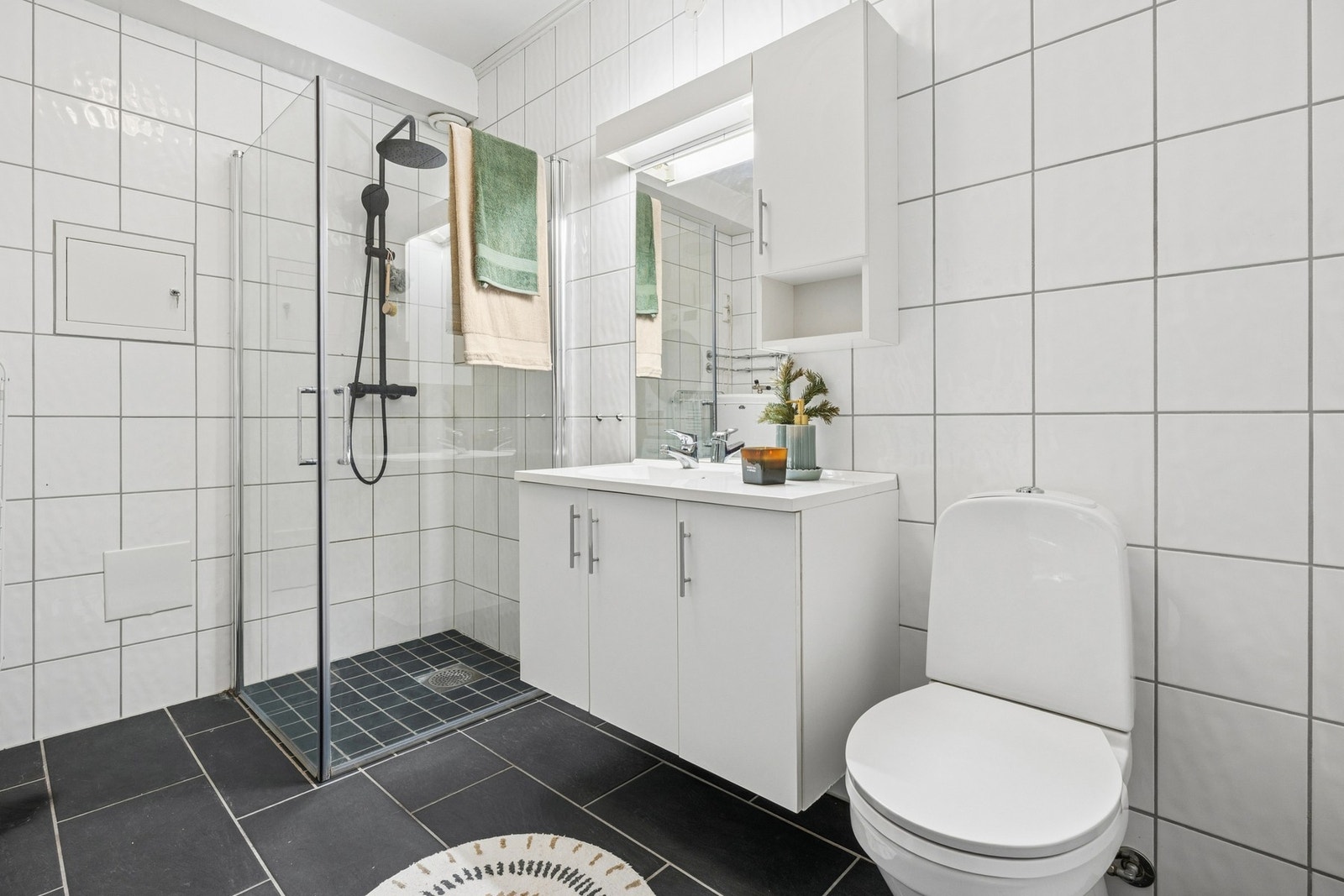 Badet i underetasjen er flislagt og inneholder dusjløsning med veggmontert armatur, servant med skap, gulvstående toalett samt opplegg for vaskemaskin og varmtvannsbereder. Galleribilde