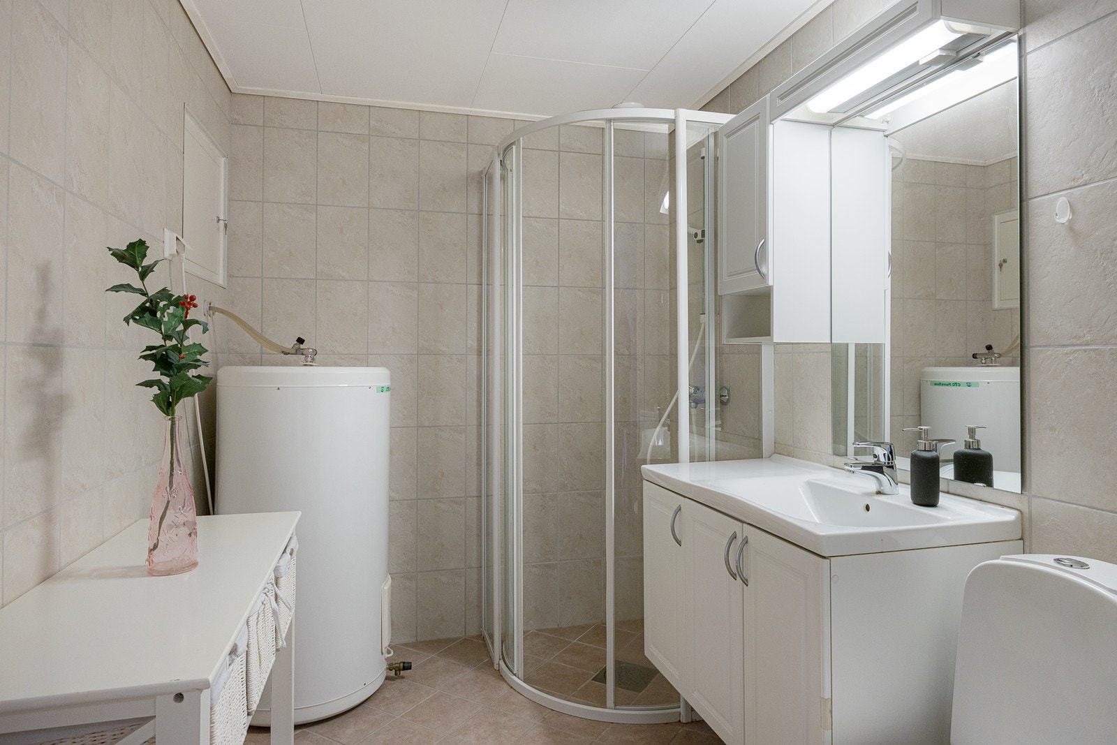 Det praktiske badet har sandfargede fliser på gulvet, lyse fliser på veggene, gulvvarme og mekanisk avtrekk. Videre er det montert speil med overlys, heldekkende servant på hvitt servantskap, og et matchende veggskap. Galleribilde