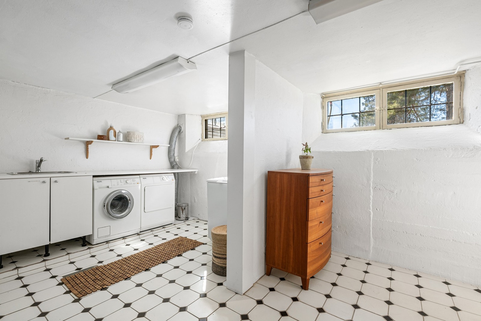 Stort vaskerom i kjeller med flislagt gulv, malte murvegger og pusset og malt himling. Det er innredning med benkeskap med glatte hvite fronter og laminat benkeplate med nedfelt skyllekar og opplegg for vaskemaskin og tørketrommel. Det er installert kjøle Galleribilde