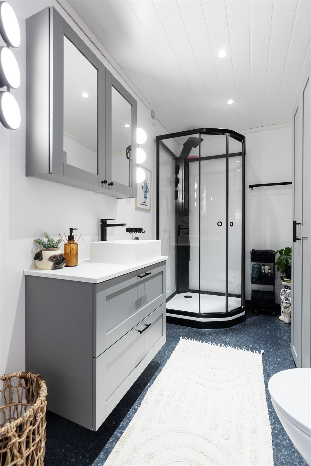 Badet har godt med oppbevaringsplass, med både servantskap, høyskap og speilskap i en delikat grå fargetone. Badet er også innredet med veggmontert toalett og dusjkabinett i stilrent design med sorte profiler og sort dusjgarnityr. Galleribilde
