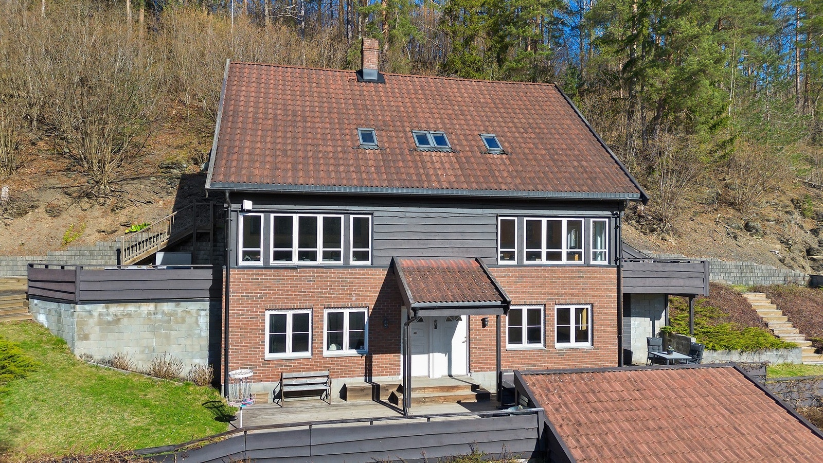 Bolig over to etasjer og loft med skråtak og flere vinduer, beliggende i skrånende terreng. Galleribilde