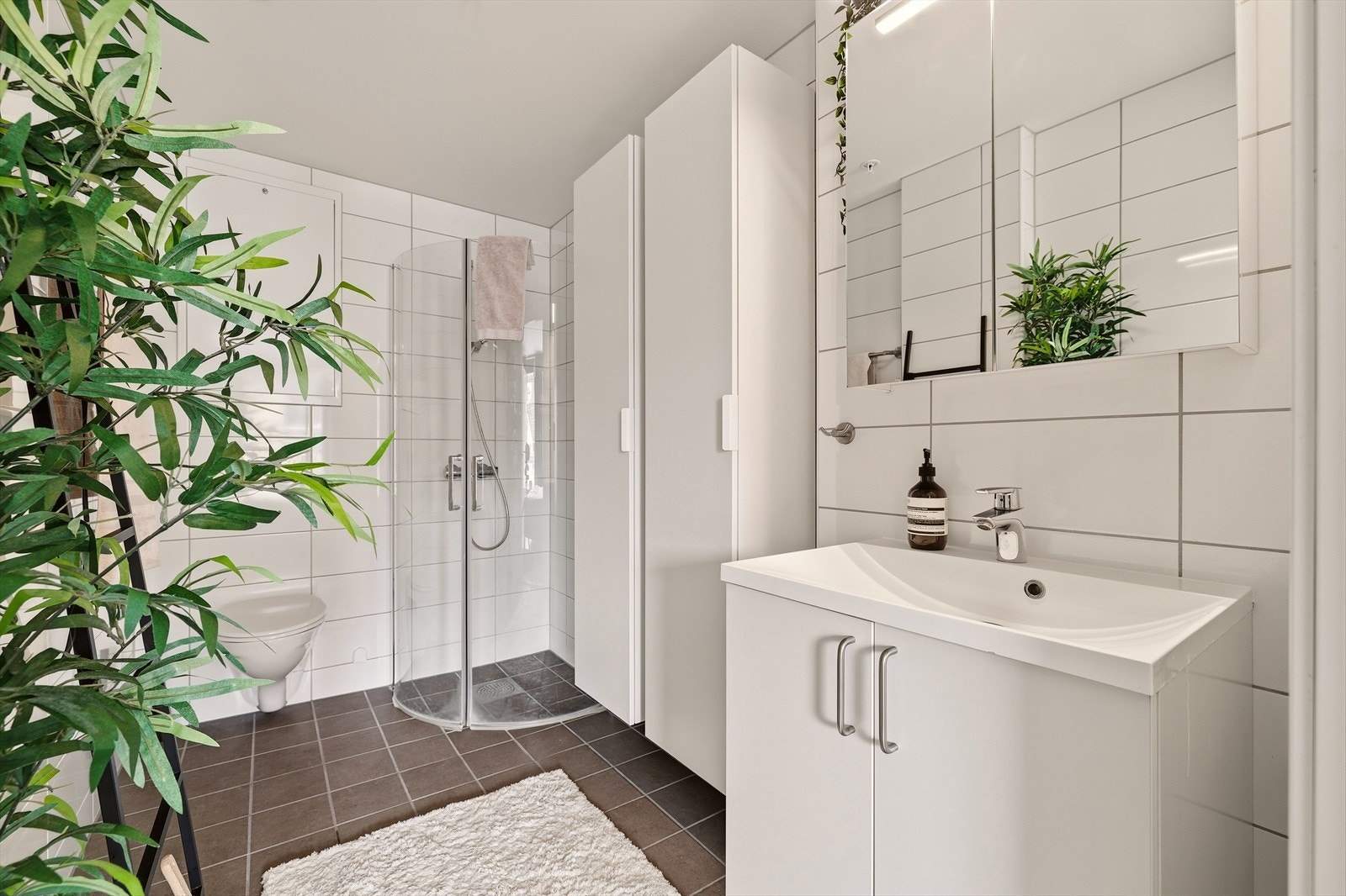 Badet inneholder servantinnredning med heldekkende servant, speilskap med vegglampe, veggskap, vegghengt toalet og dusj med innfellbare dører i herdet glass. Her er vannbåren varme i gulv og avtrekksventil i himling. Galleribilde