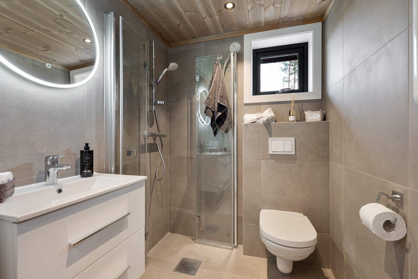 Badet har vegghengt toalett, dusjhjørne med foldbare glassdører og heldekkende servant over hvit skuffeseksjon. Galleribilde