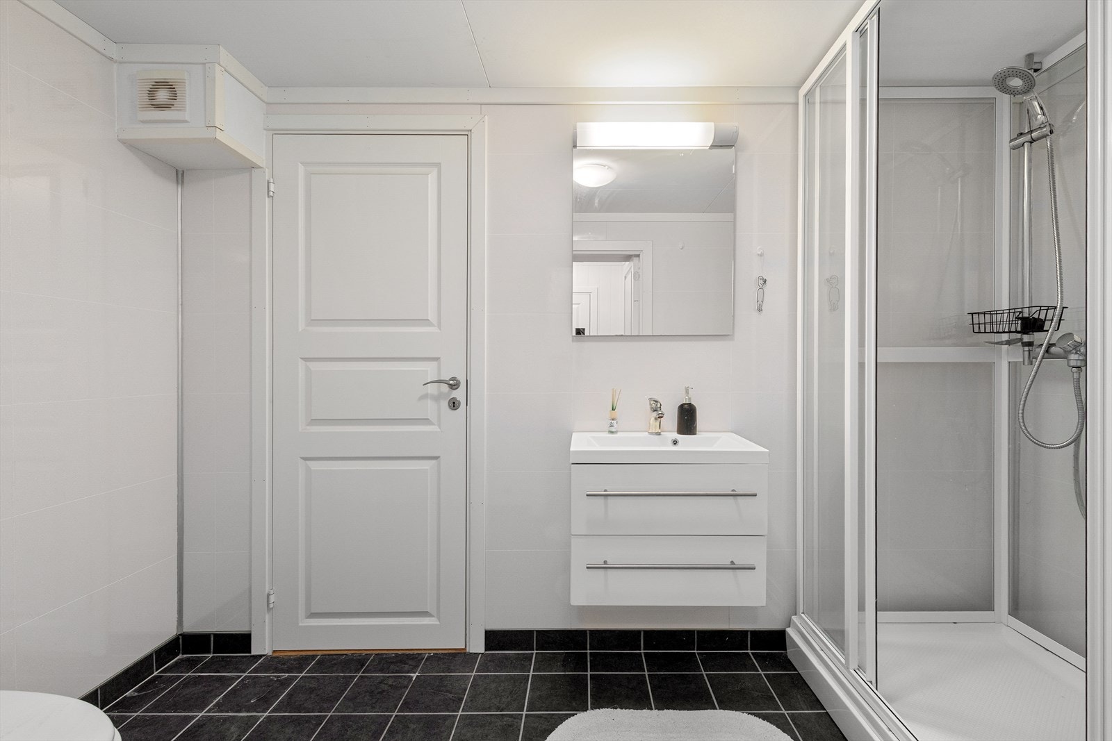 Bad med fliser på gulv, baderomsplater på vegger og kvite himlingsplater. Badet er utstyrt med dusjkabinett, toalett og servant. Galleribilde