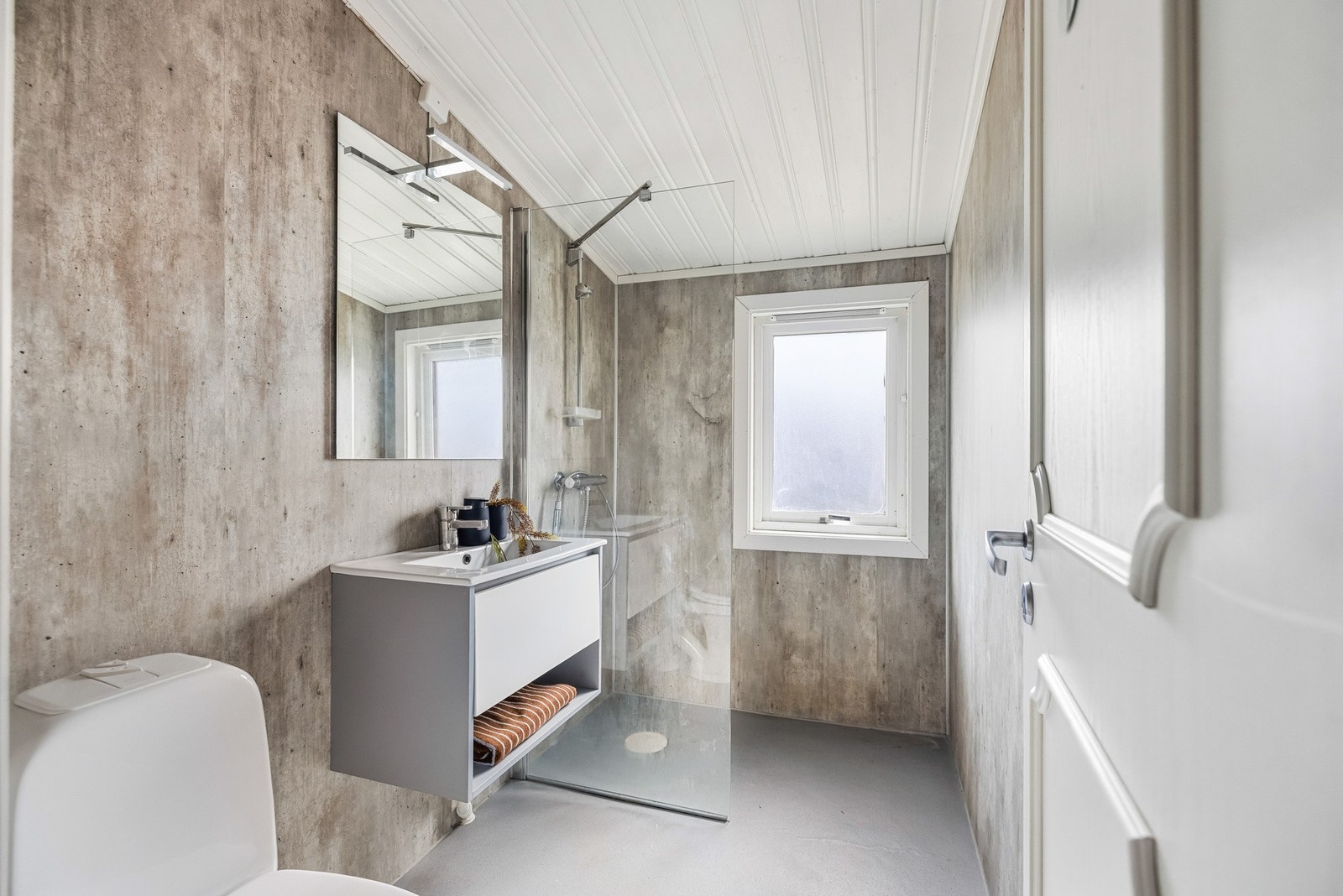 Badet er innredet med baderomsplater på vegger og belegg på gulv, med dusjhjørne, gulvmontert toalett og baderomsinnredning med heldekkende servant og speil på vegg. Galleribilde