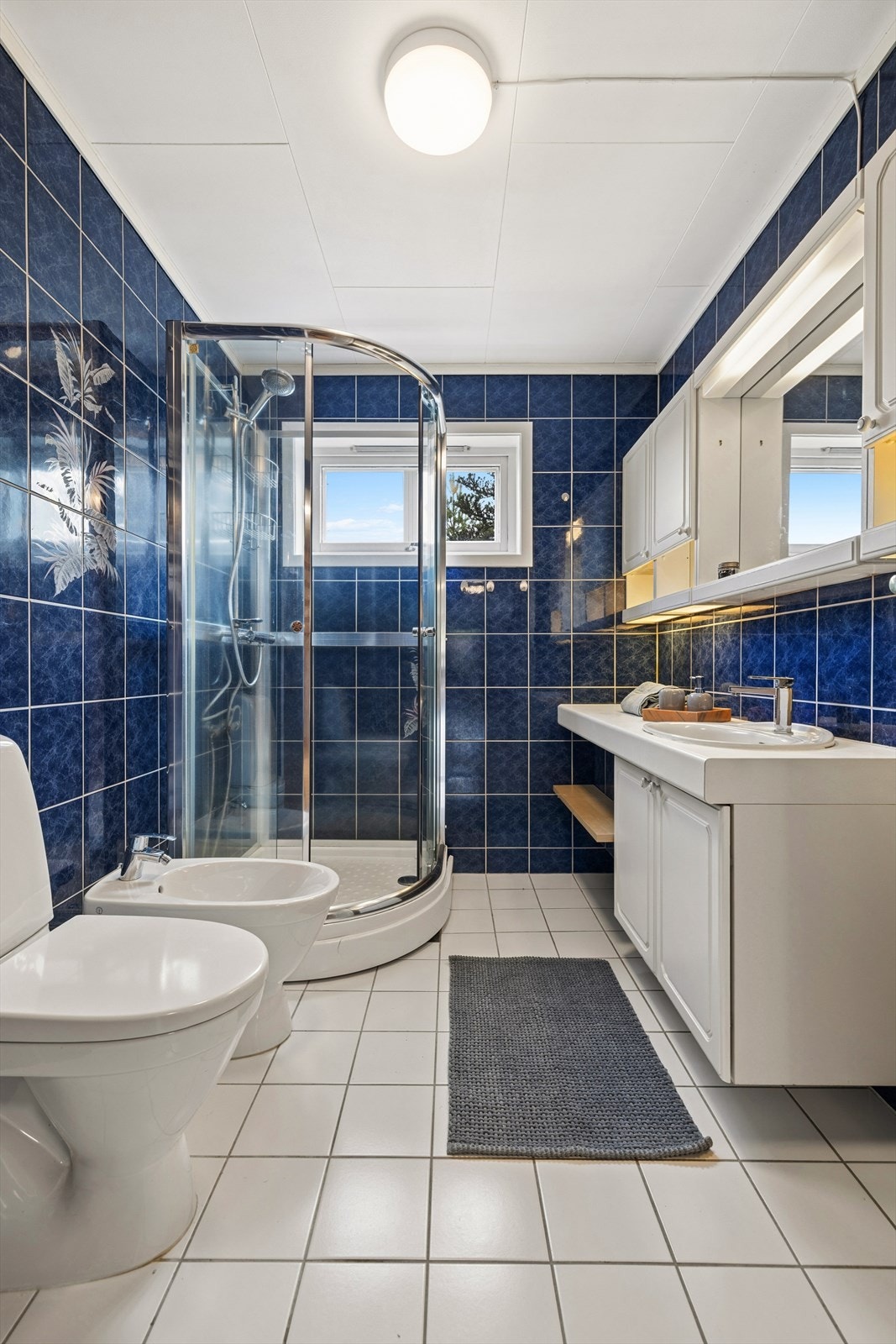 Badet er fra byggeår og er innredet med hvite fliser på gulvet og blå fliser på vegg. I gulvet har man også varmekabler for ekstra komfort. Badet er ellers utstyrt med dusjkabinett, toalett, bidé og baderomsinnredning med nedfelt servant. Galleribilde