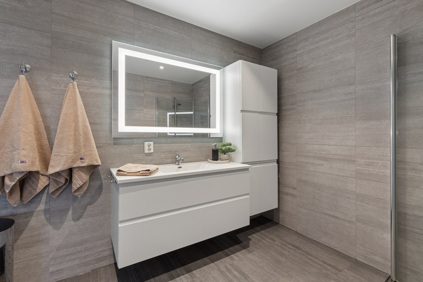 Badet har moderne og tidløse løsninger med flislagte vegger og gulv, samt elektriske varmekabler. Rommet er innredet med nedfelt servant over skuffer som gir praktisk oppbevaring, og speil med integrert belysning som skaper et stilrent uttrykk. Galleribilde