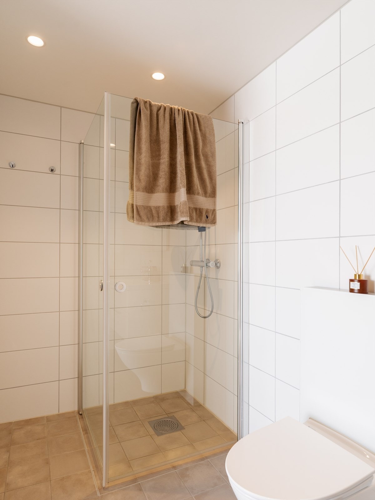 Her er det hvite fliser på veggene, downlights i himlingen og sandfargede fliser på gulv med elektrisk gulvvarme. Badet har et vegghengt toalett og et dusjhjørne med innfellbare glassdører. Galleribilde