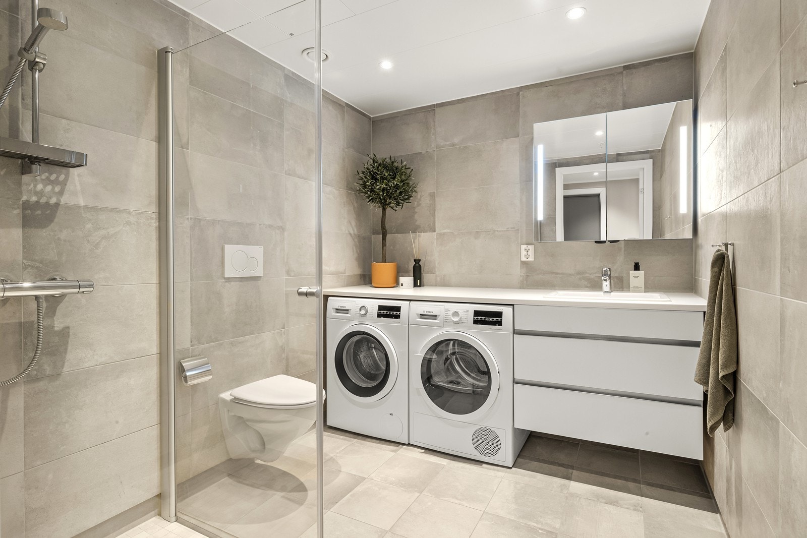 Badet er flislagt og har vannbåren gulvvarme. Romslig og praktisk løst med heldekkende benkeplate ut fra baderomsinnredningen der vaskemaskin og tørketrommel er plassert under. Galleribilde