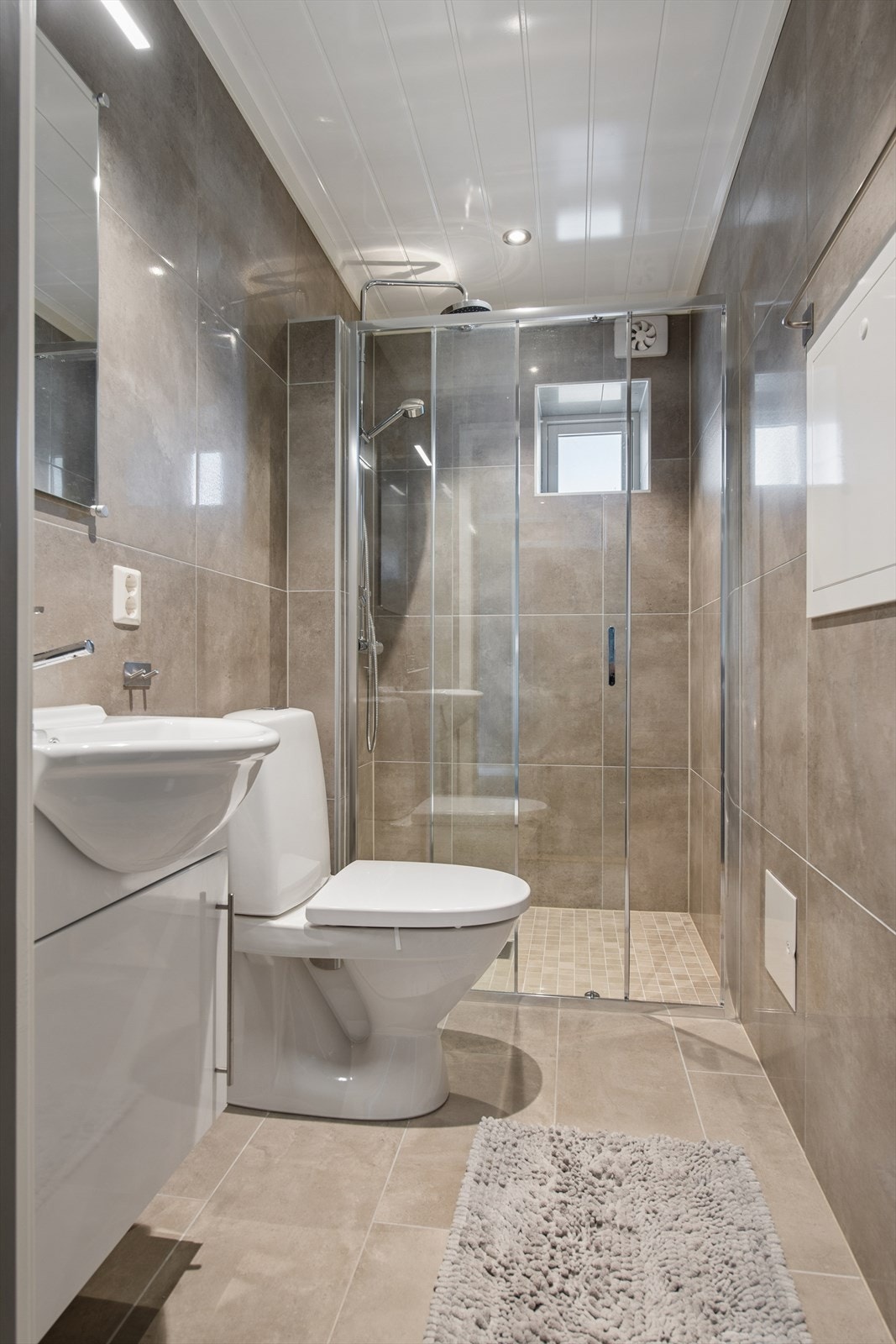 Badet fra 2017 har fliser på gulv og vegger. Rommet er utstyrt med servant med underskap, speil med belysning, gulvmontert toalett og en dusj med skyvedør. Galleribilde