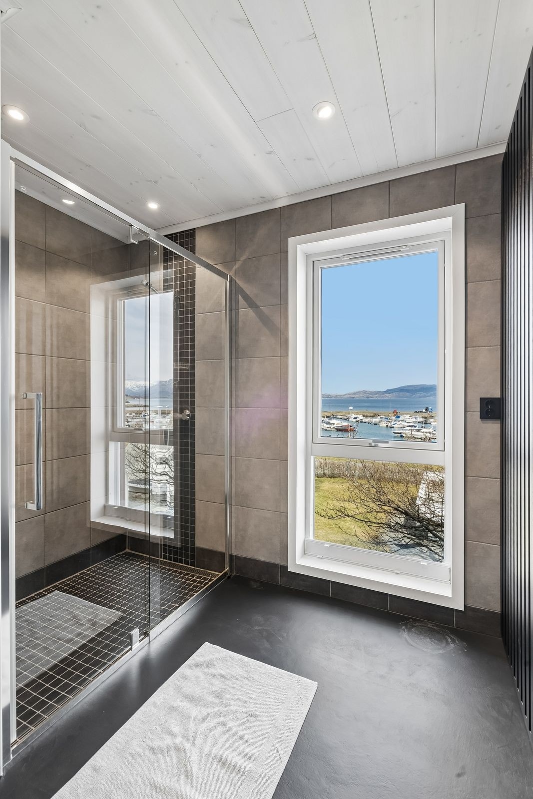 Badet har store flislagte overflater, romslig dusjnisje med glassvegger og stilren baderomsinnredning. Til venstre i bildet ser man det praktiske avlukket som rommer vaskemaskin og tørketrommel. Galleribilde