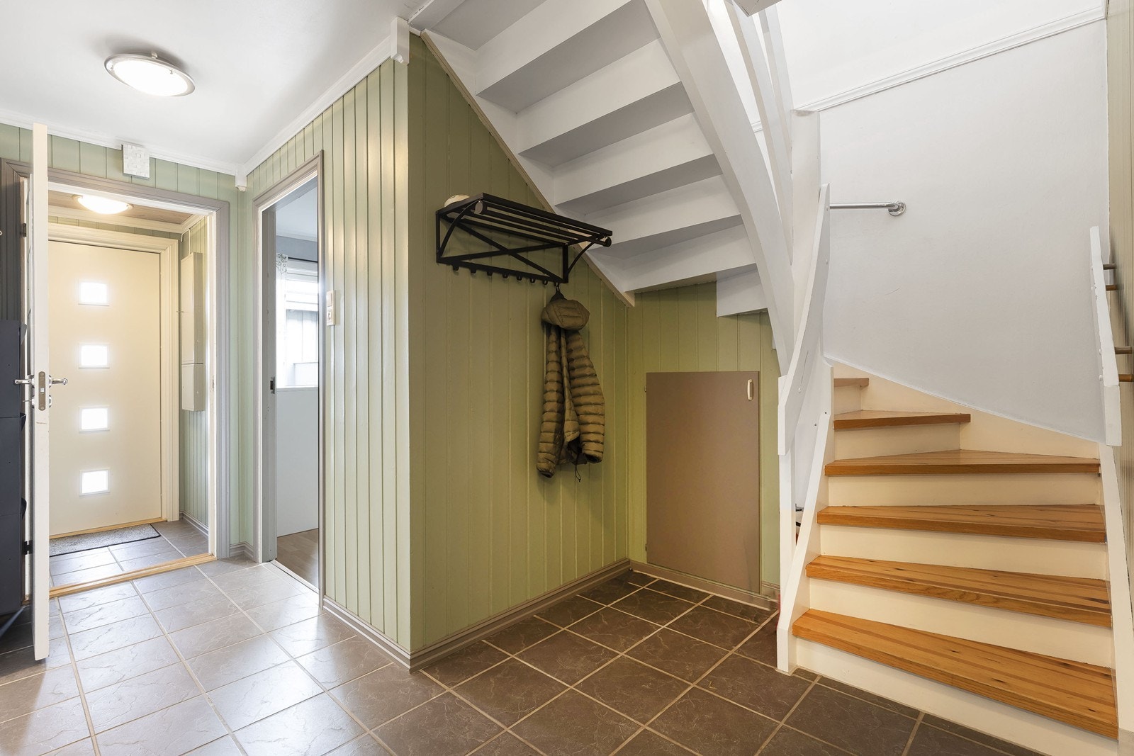 Hall med flislagt gulv og malte panelvegger. Oppbevaringskott under trappa. Galleribilde