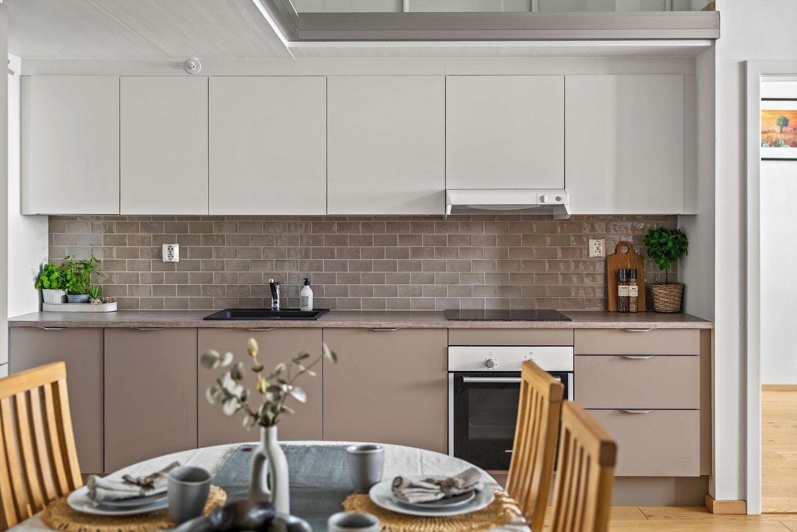 Stue og kjøkken er i åpen løsning, noe som gir en sosial hovedsone. Kjøkkenet fra HTH har en moderne og helhetlig utforming, med integrerte hvitevarer og en stilren kombinasjon av beige og hvite fronter. Galleribilde