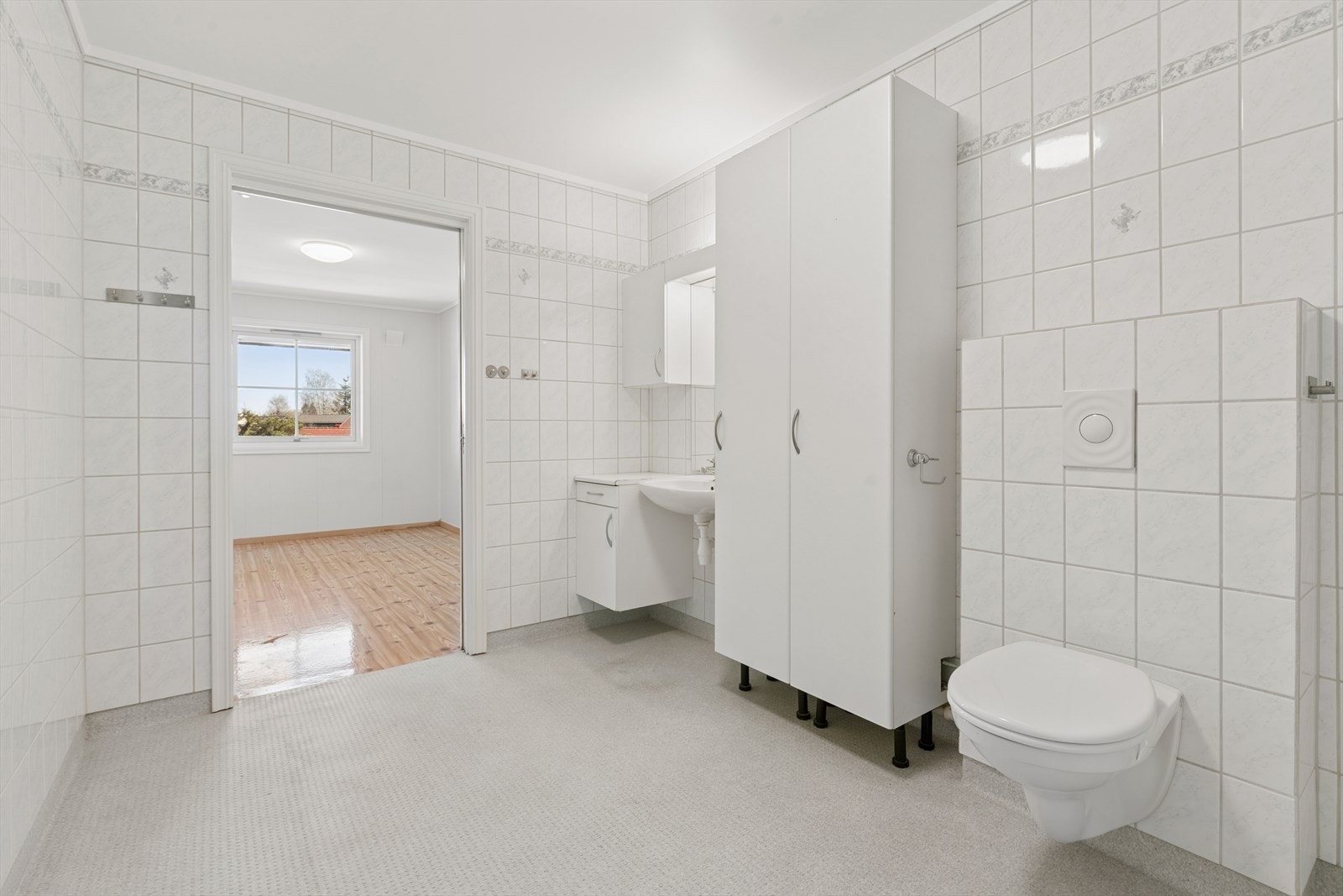 Baderomsinnredning med høyskap og sideskap med hvite glatte fronter. Rommet har servant, toalett, dusjvegger/hjørne og opplegg for vaskemaskin. Galleribilde