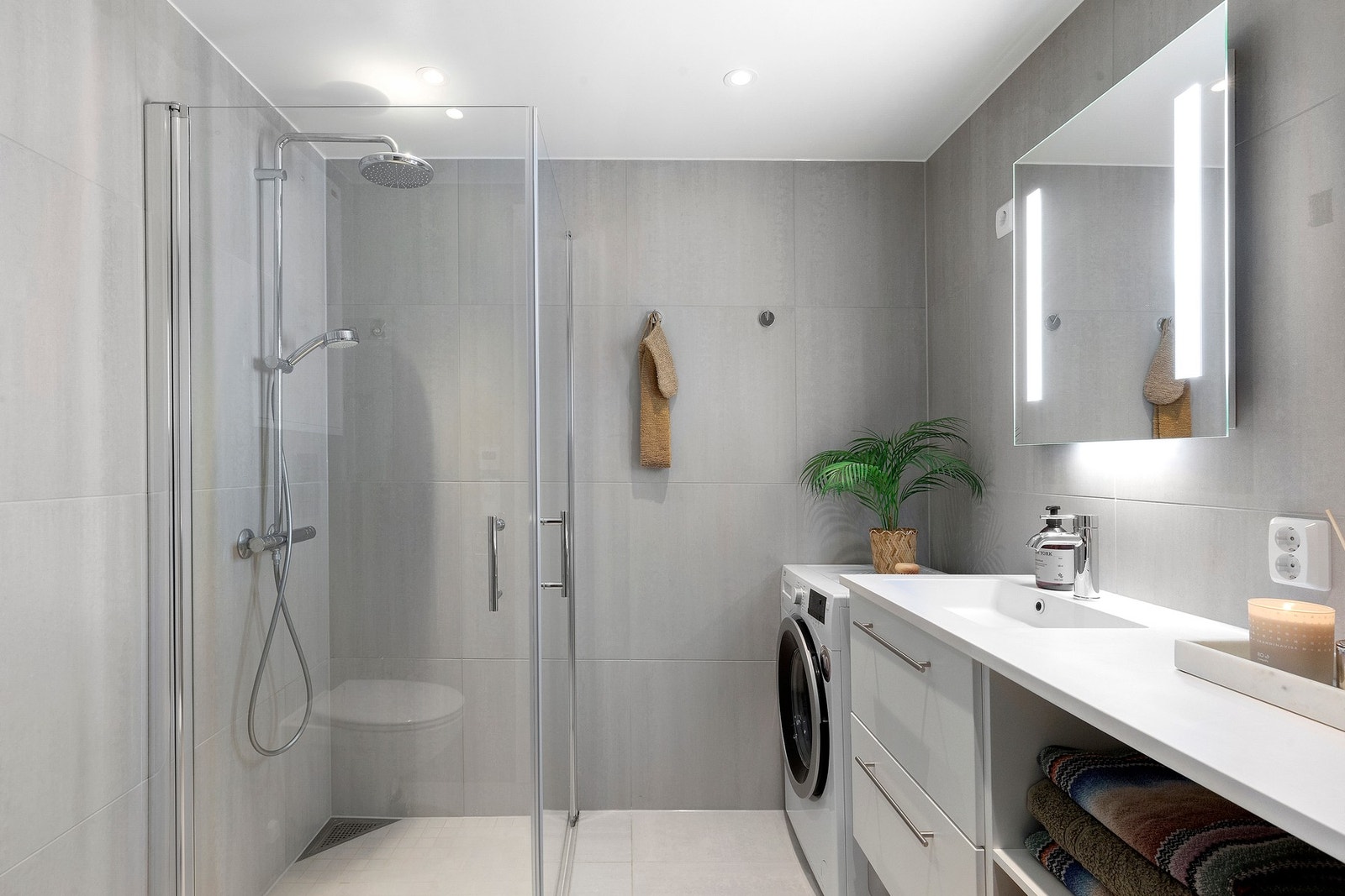 Badet på 4,2 kvm har fine store fliser, innfellbare dusjdører, regndusj, praktiske skuffer og hyller til oppbevaring, opplegg til vaskemaskin og moderne speil med integrert lys Galleribilde