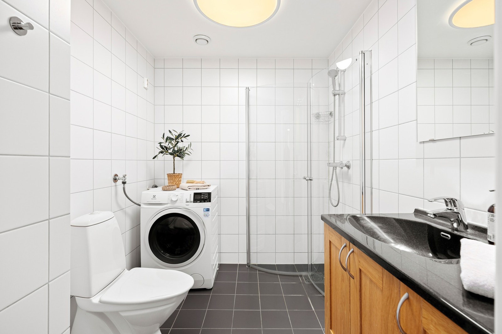 Badet er innredet med en heldekkende servant montert på en vegghengt skapseksjon, speil og vegghengt skap. Videre er rommet utstyrt med frittstående toalett, dusjhjørne og opplegg for vaskemaskin Galleribilde