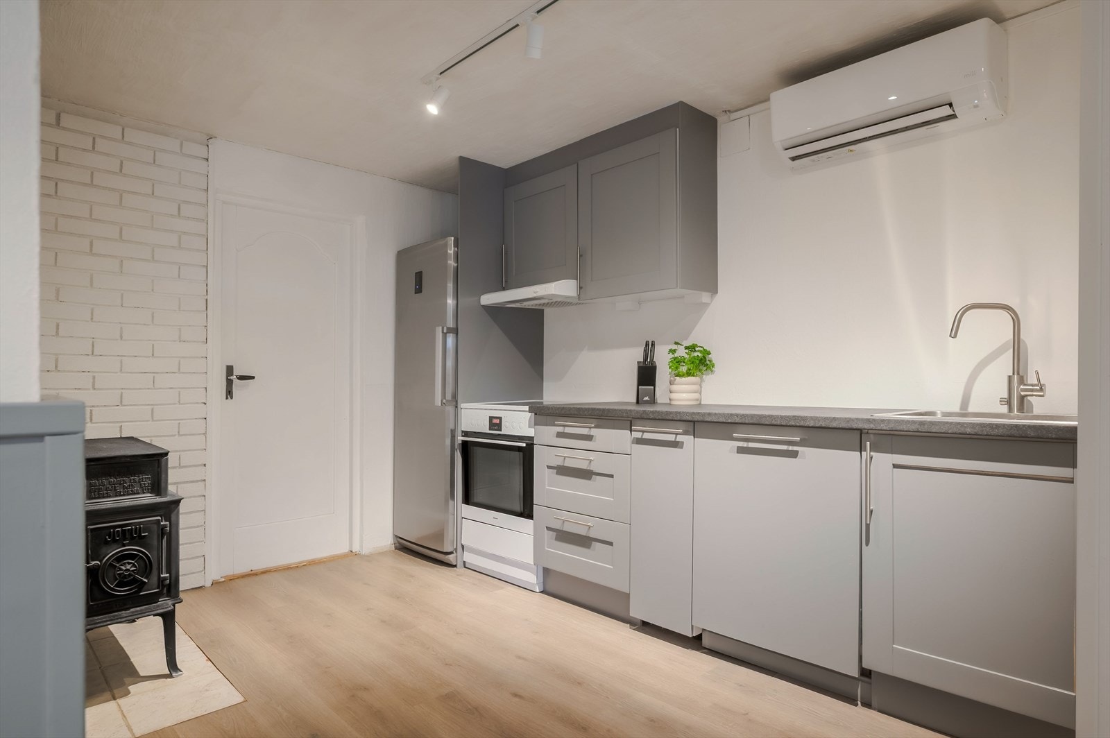 Moderne kjøkkeninnredning med grå fronter og laminert benkeplate med nedfelt oppvaskkum. Kjøkkenet har både vedovn og nyere varmepumpe. Galleribilde