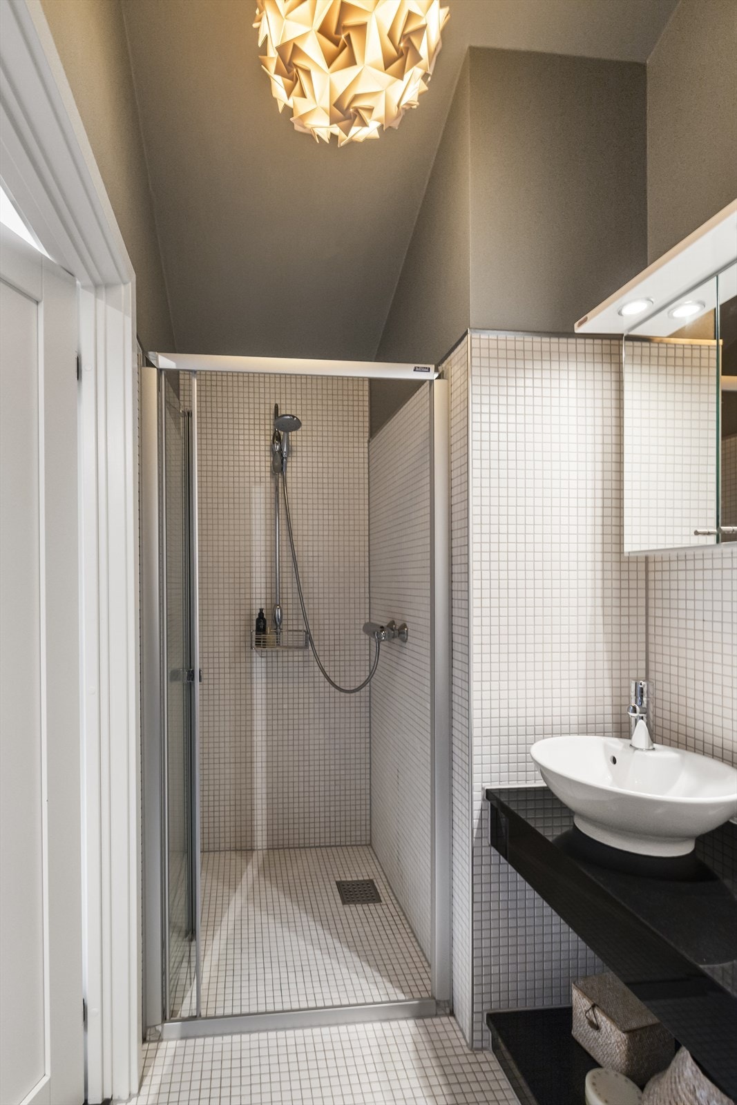 Badet har mosaikkflis på overflatene og vannbåren gulvvarme. Det er speilskap, belysning og toppmontert servant på plassbygget møblement. Badet har vegghengt toalett og dusjnisje med foldedører. Galleribilde