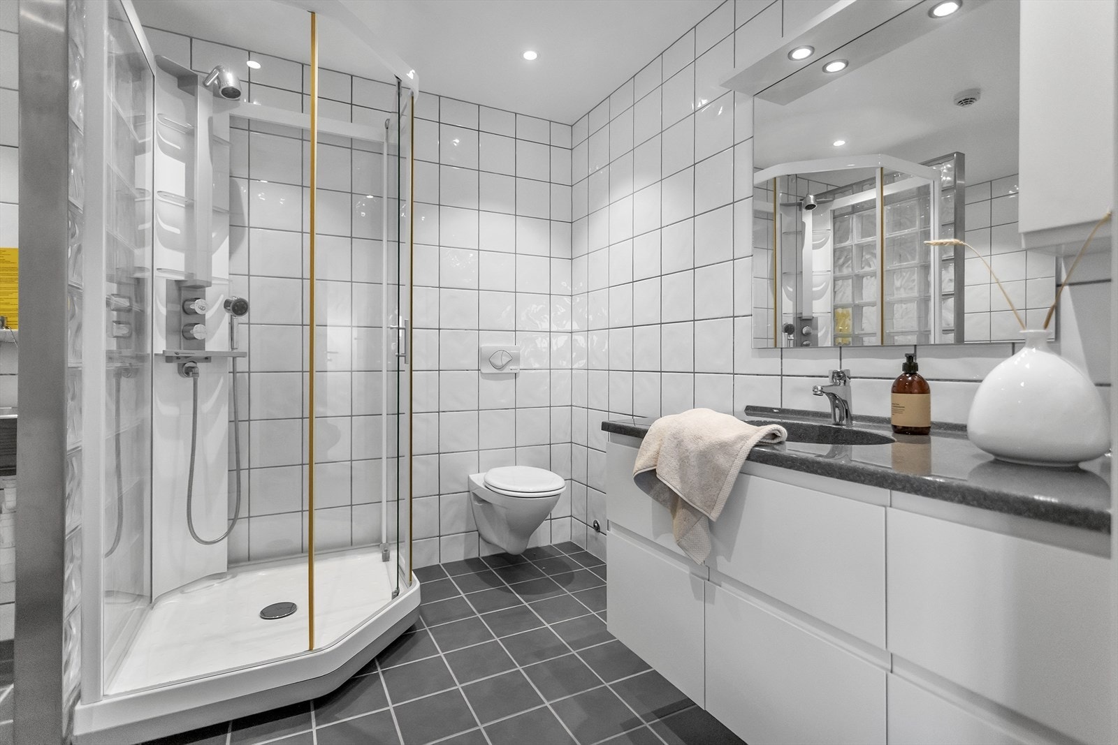 Badet er stilrent og helfliset, rommet er utstyrt med et praktisk dusjkabinett, servant med både overskap og underskuffer for smart oppbevaring, samt vegghengt toalett som gir et moderne og ryddig uttrykk. Galleribilde