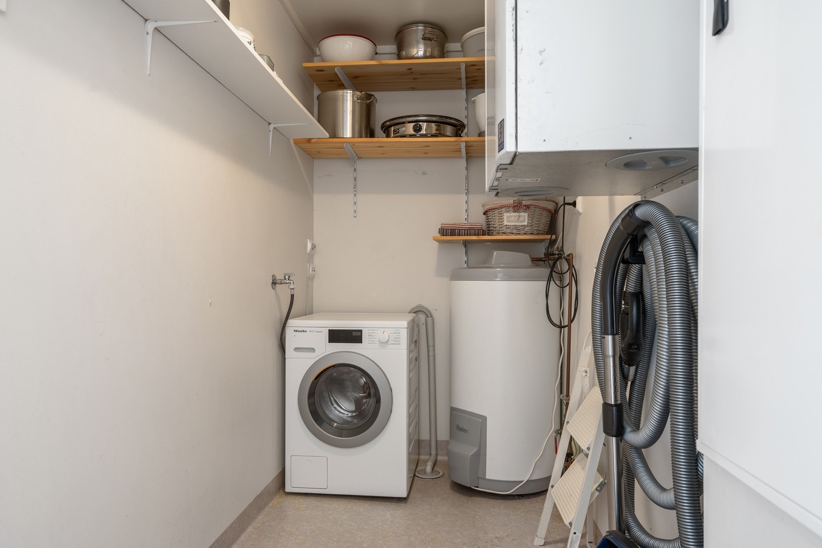 Praktisk bod/vaskerom innredet med malte plater på vegger, fabrikkmalte takplater og vinylbelegg på gulvet. Rommet har plastsluk og opplegg for vaskemaskin. Galleribilde