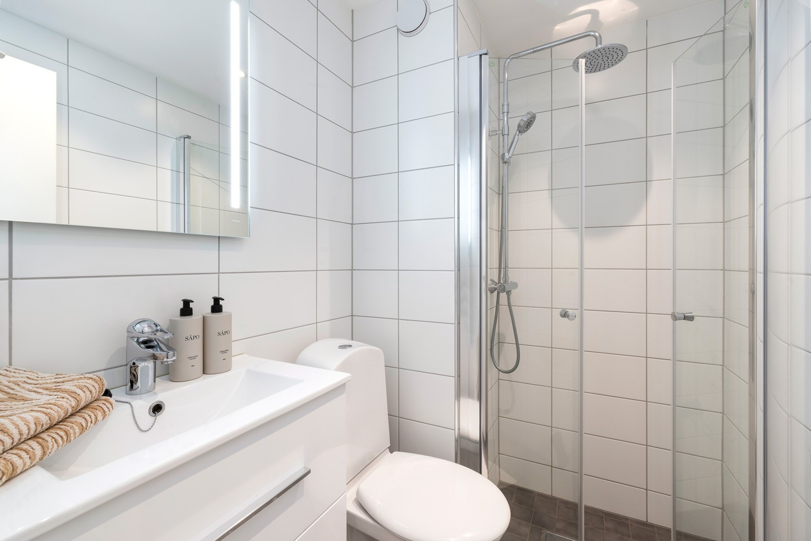 Badet er flislagt og innredet med servant, speil med lys, wc og dusjhjørne Galleribilde