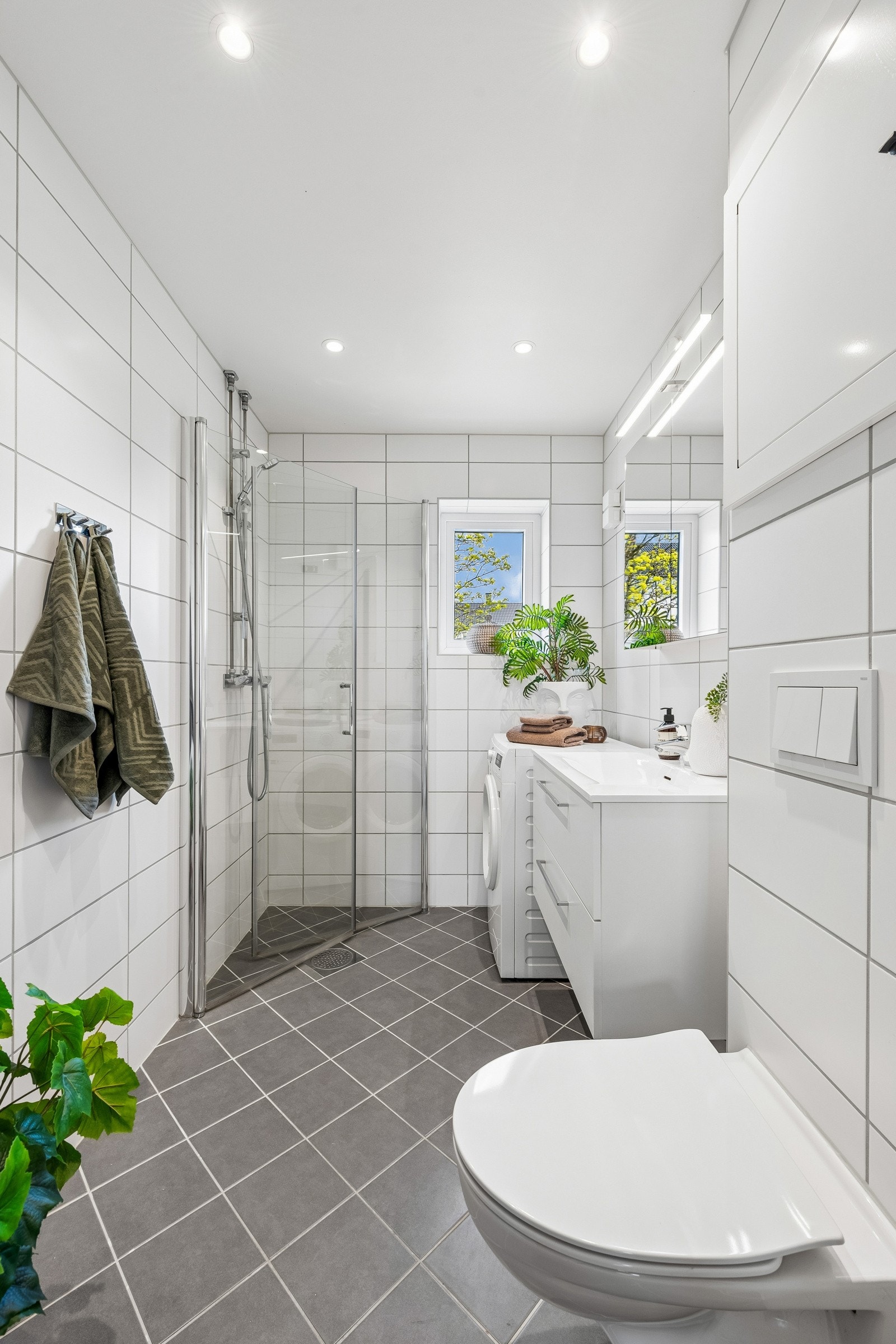 Innbydende baderom med dusj med inn/- utadslående dusjvegger, servant med underskap, overskap med speilfronter og overlys. Veggmontert klosett og opplegg for vaskemaskin. Kombinert vaskemaskin/tørketrommel medfølger. Galleribilde
