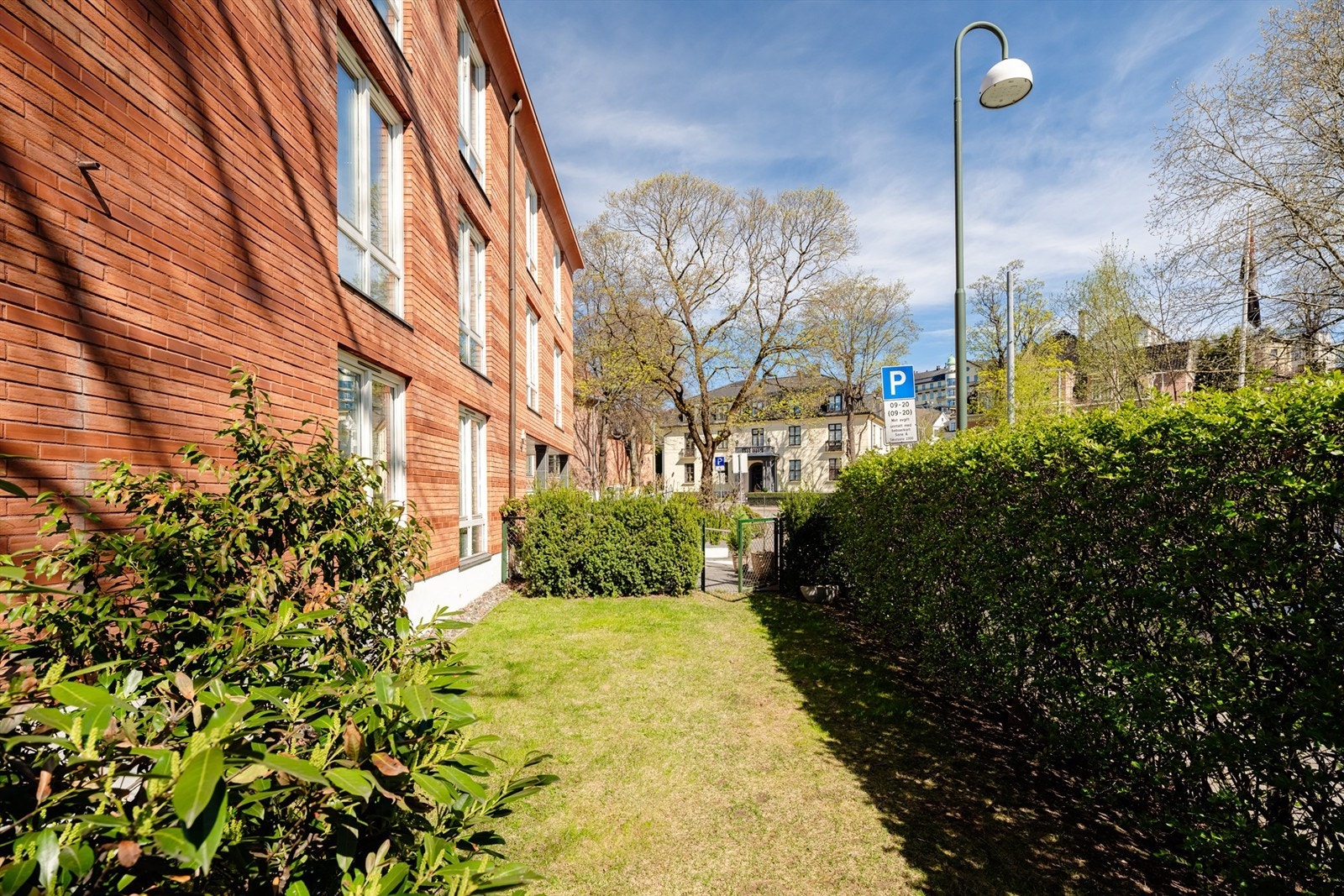 Pent opparbeidet plen med busker og blomster rundt bygningen. Hellelagt inngangsparti med bred trapp og asfaltert nedkjørsel til garasjeanlegg. Galleribilde