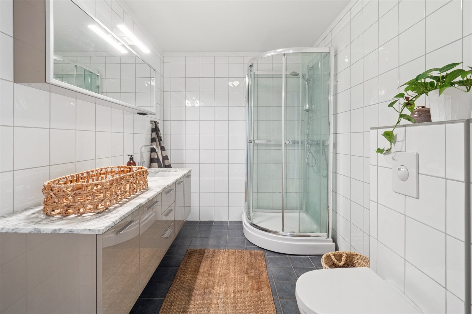 Pent bad med fliser på vegger og gulv. Badet har servant med skap og skuffeseksjon, speil med tilhørende belysning, dusjkabinett og vegghengt wc. Galleribilde