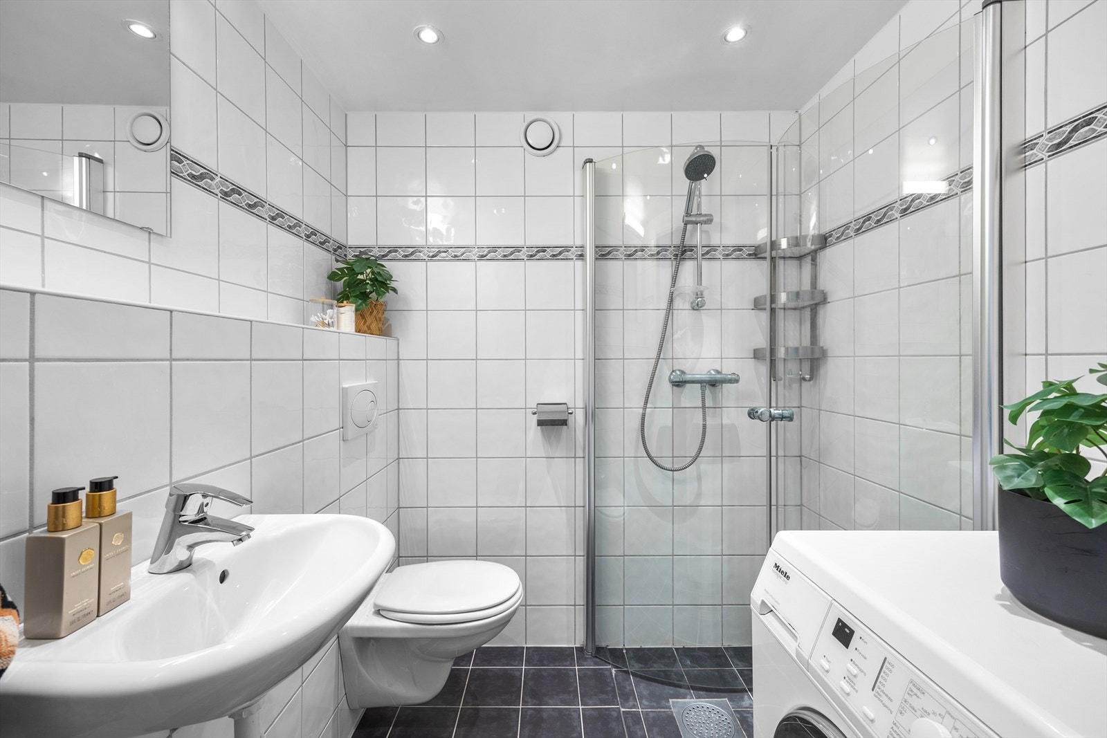 Badet har flislagt gulv med elektriske varmekabler som gir god komfort. Rommet er innredet med servant med speil og belysning over, veggmontert toalett samt dusjhjørne med glassvegger. Galleribilde