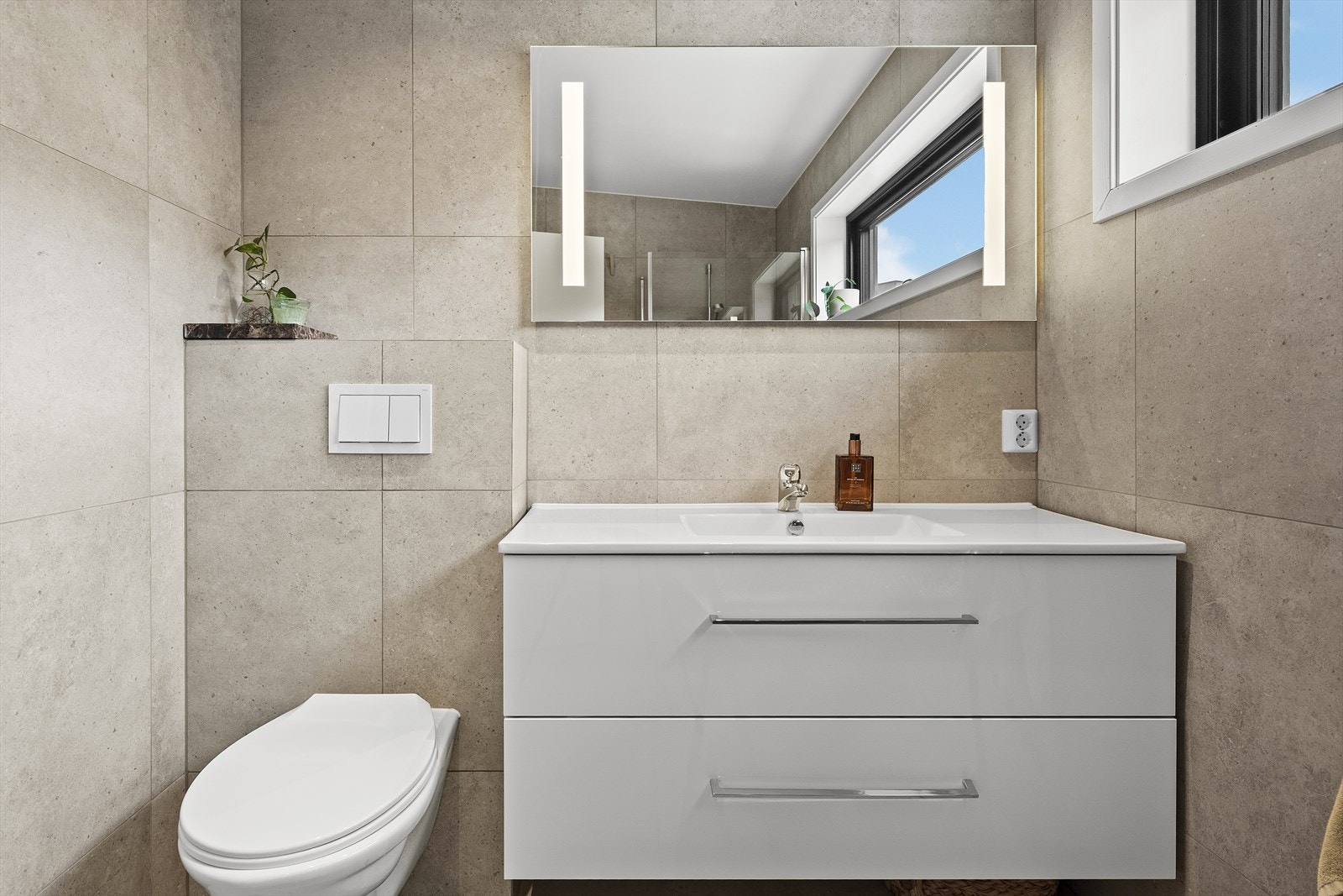 Baderommet er utstyrt med heldekkende servant over skuffeinnredning, overliggende speil med belysning, vegghengt toalett, samt dusjhjørne med innfellbare dusjvegger Galleribilde