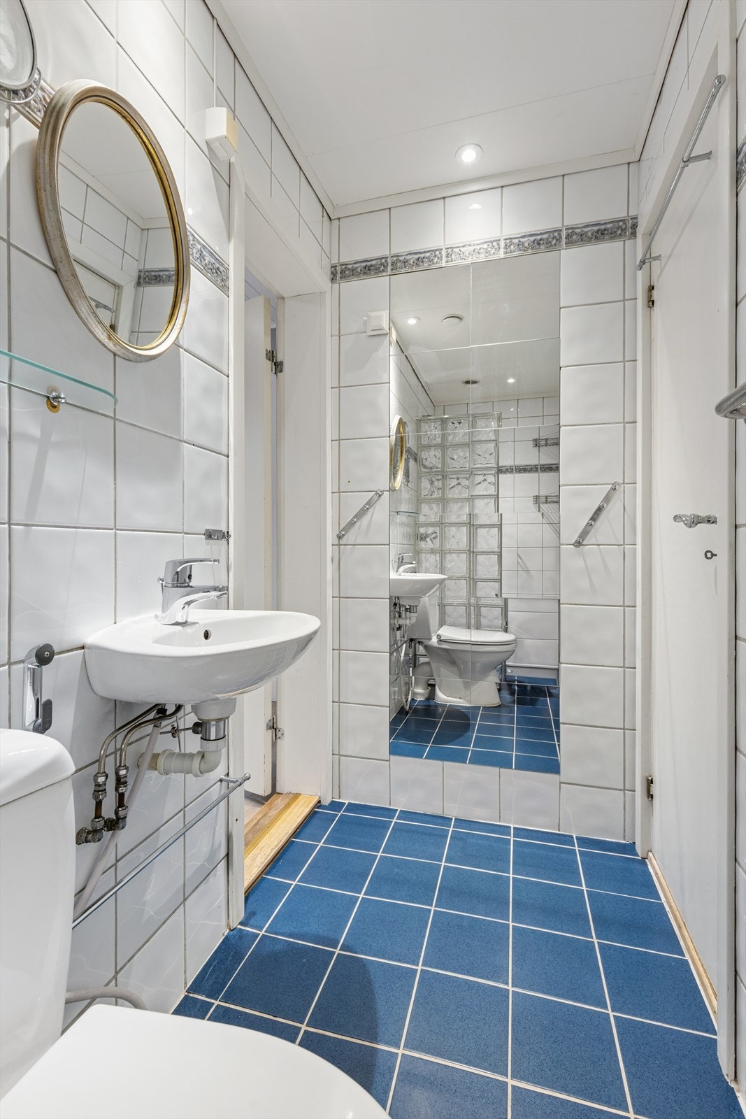 Bad med flislagte vegger i hvitt, blått flisgulv, servant med speil over, toalett og speilvegg som reflekterer dusjsonen med glassbyggestein. Galleribilde