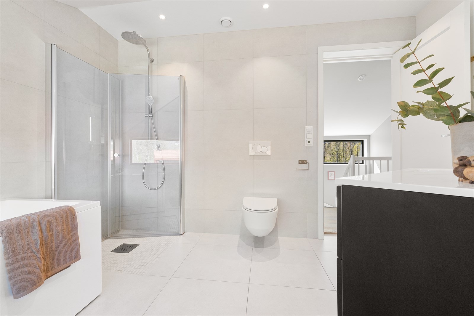 Badet er utstyrt med badekar, dusjhjørne med innfellbare glassdører og veggmontert toalett. Moderne baderomsinnredning med skuffer, servant med ettgreps armatur og speil med lys. Galleribilde